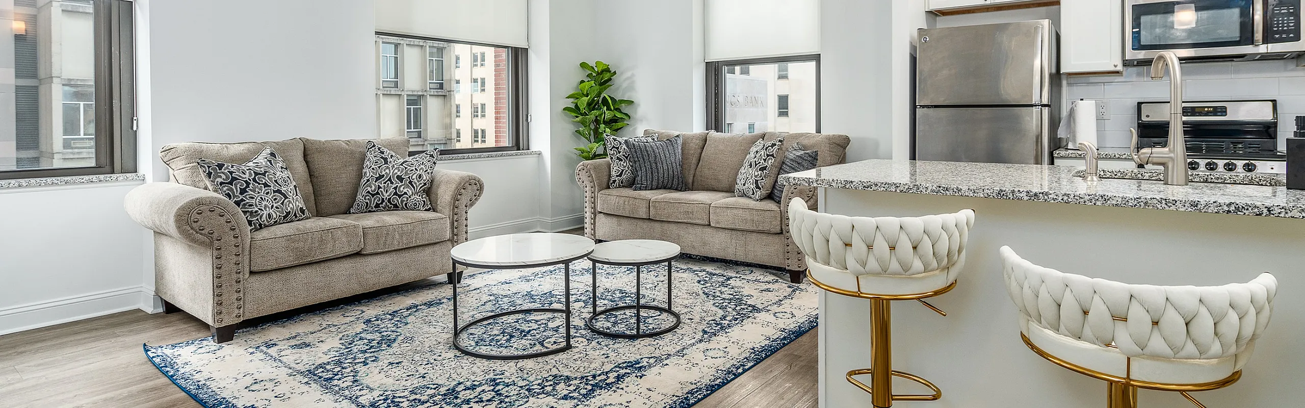 Chic open-concept living area with modern kitchen, featuring sleek stainless steel appliances, elegant island seating, and cozy sofas on stylish rug.