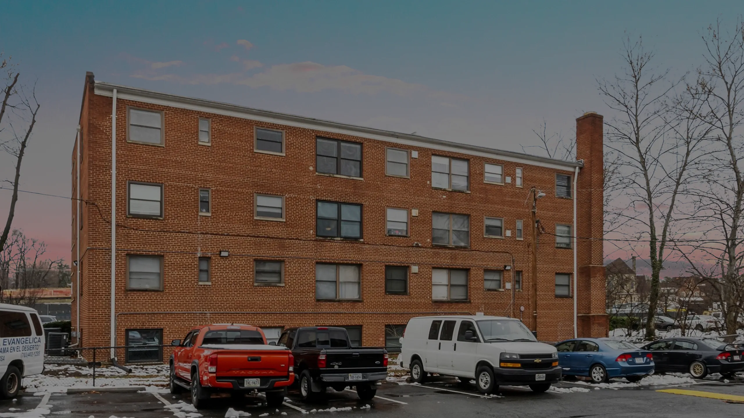 Spacious three-story brick apartment building with parking area set against a serene dusk sky.