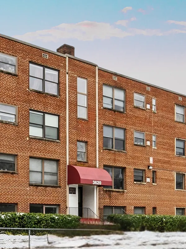 Red brick apartment building with multiple windows and a welcoming entrance canopy, set against a colorful sky.