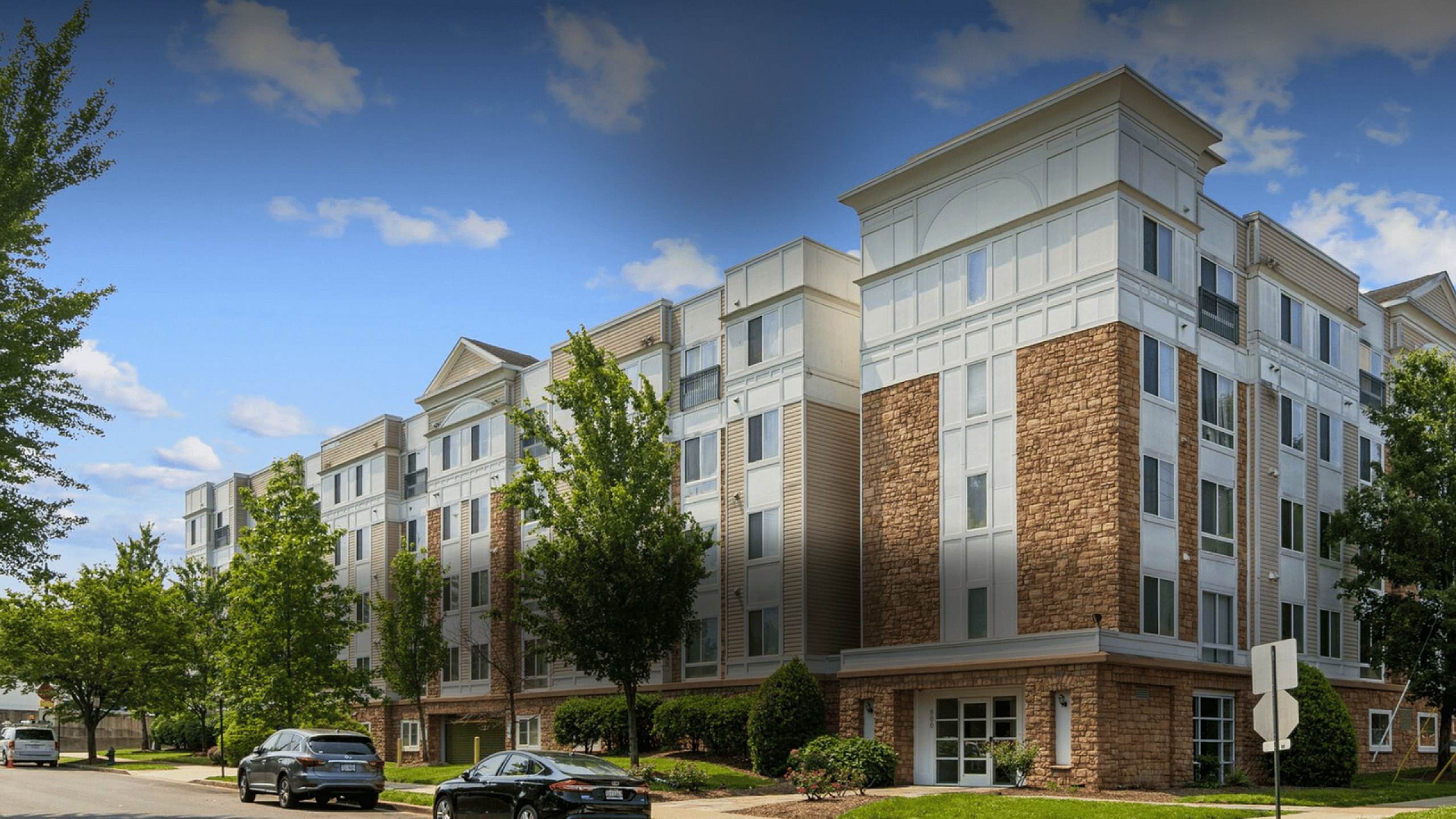 Elegant modern apartment building with classic stone facade, surrounded by lush trees and clear blue skies.