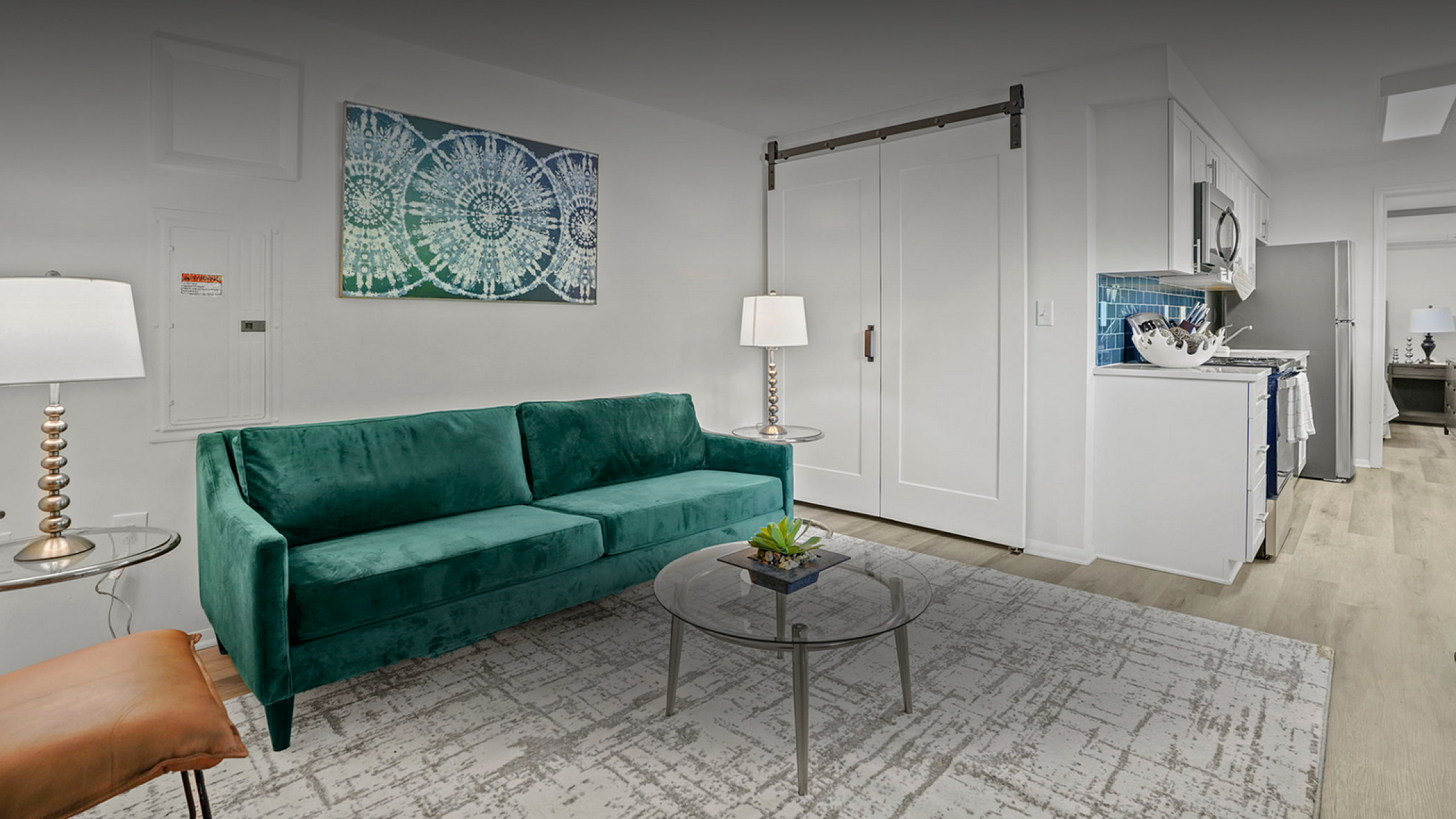 Chic apartment interior with a plush green sofa, stylish glass coffee table, and modern white kitchen featuring stainless steel appliances.