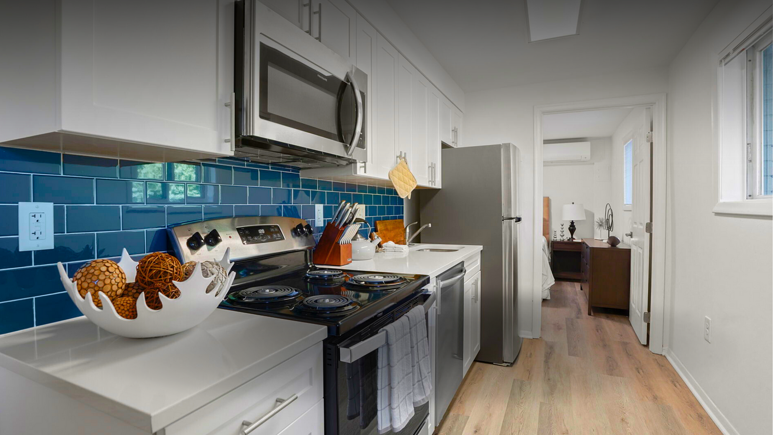 Chic apartment kitchen with modern stainless steel appliances, glossy blue tile backsplash, and sleek white cabinetry, leading to a cozy bedroom.