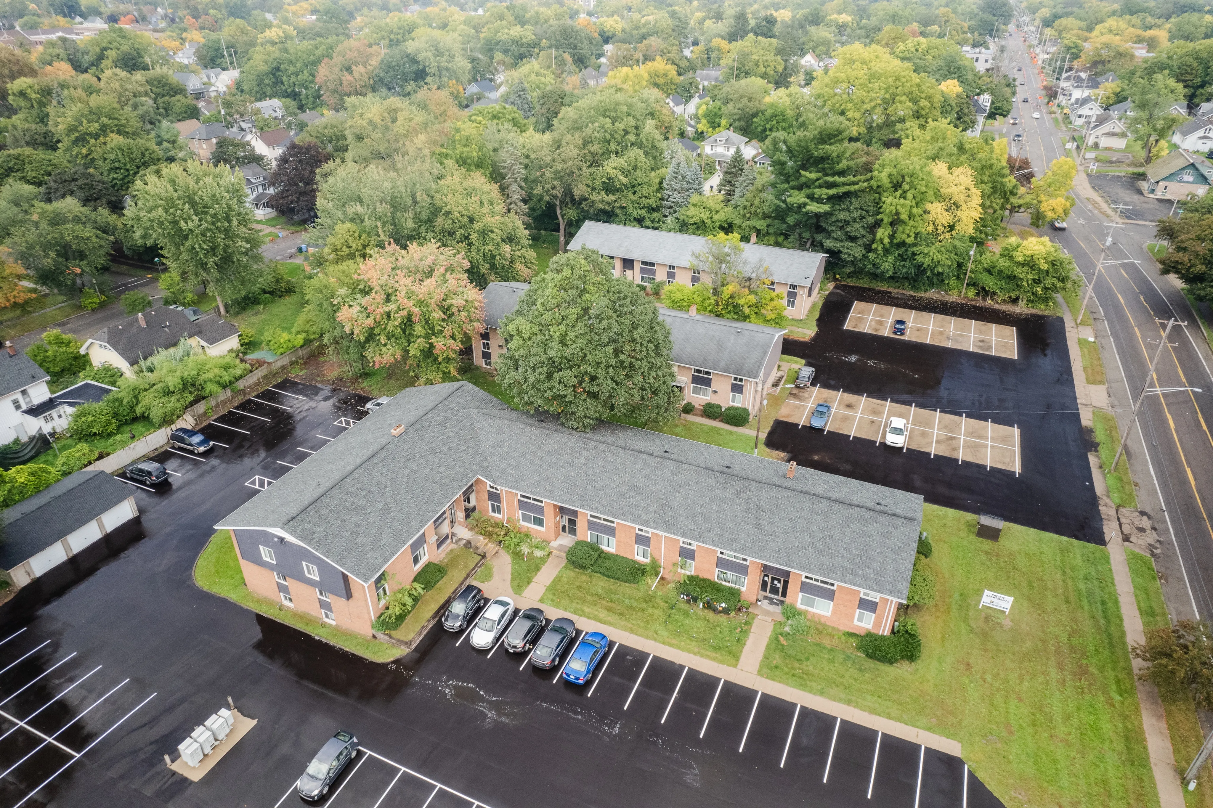 Charming aerial view of a residential apartment complex surrounded by lush greenery, showcasing ample parking and nearby tree-lined streets.