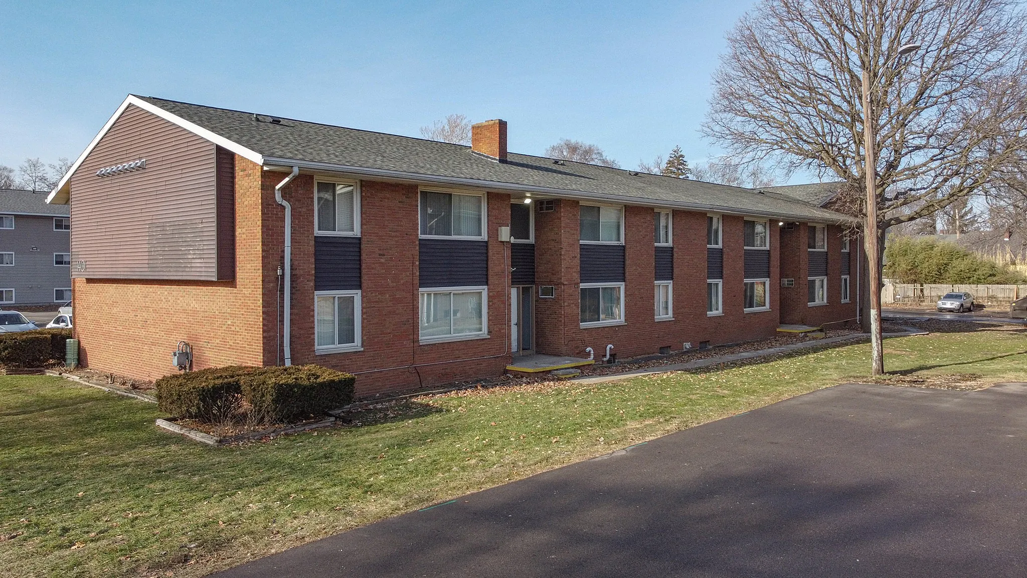 Charming two-story brick apartment building with large windows, surrounded by lush greenery and ample parking space.