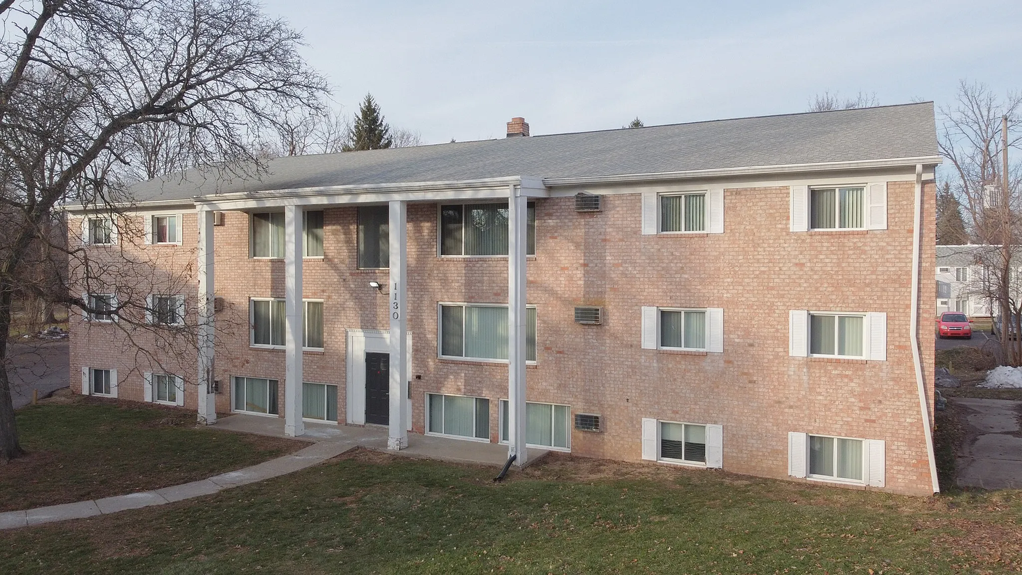Charming mid-century brick apartment building with white columns, surrounded by mature trees and a well-maintained lawn.