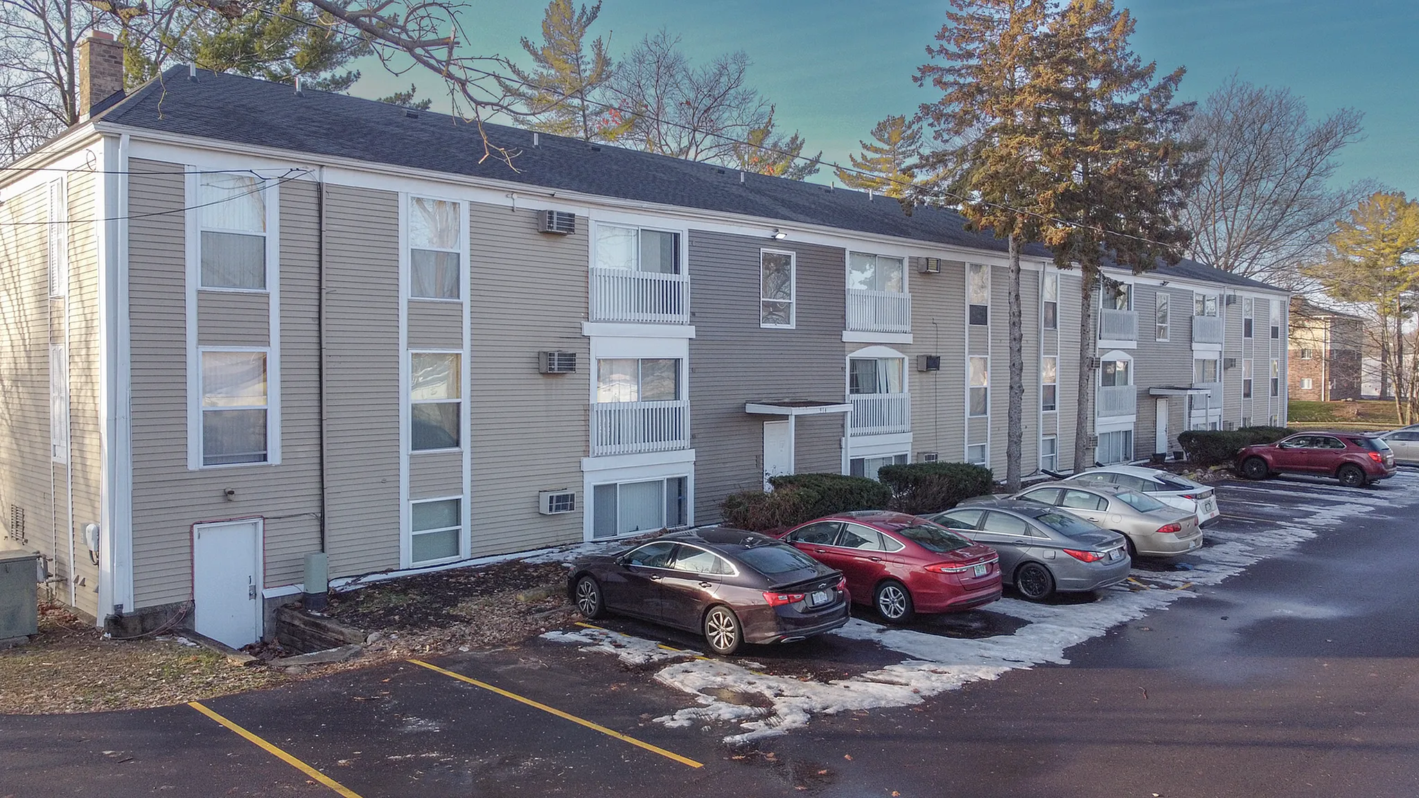 Charming three-story apartment building with modern siding and ample parking space, surrounded by lush trees under a clear blue sky.