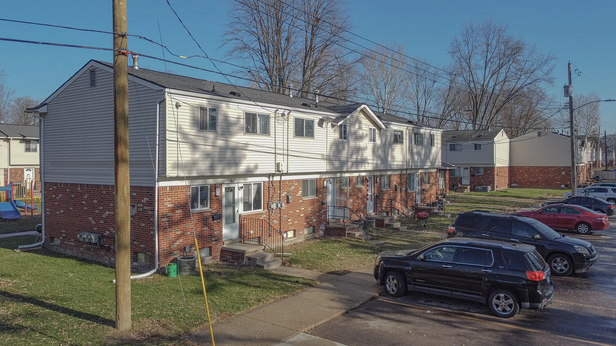 Charming row of townhouses with brick and siding exterior, set against a backdrop of mature trees, offering ample parking and a family-friendly atmosphere.