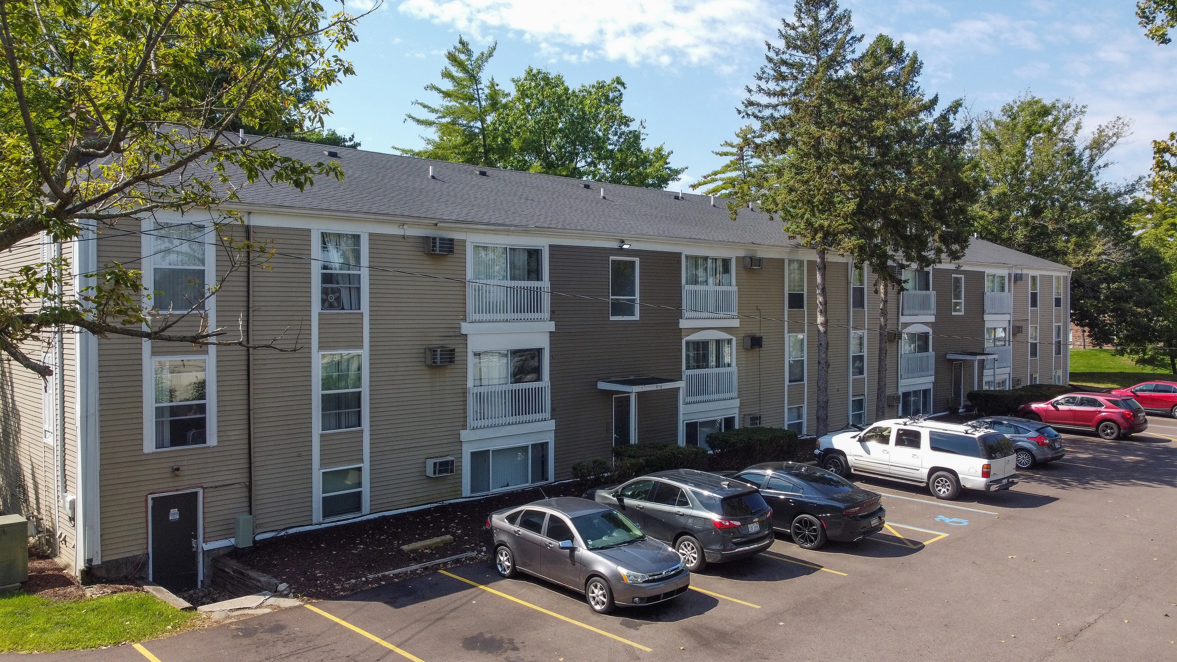 Modern three-story apartment building surrounded by lush green trees, featuring ample parking and contemporary architecture.