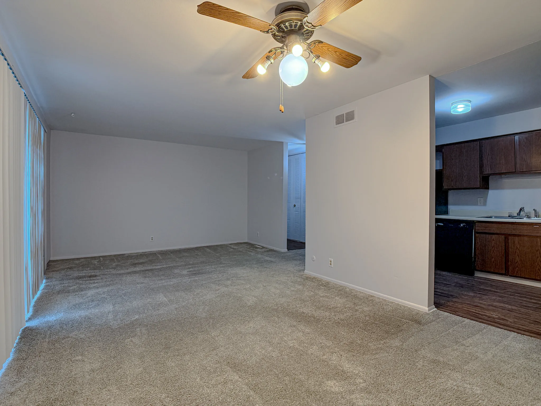 Spacious living area with plush carpet and ceiling fan, adjacent to a modern kitchen with wooden cabinets.