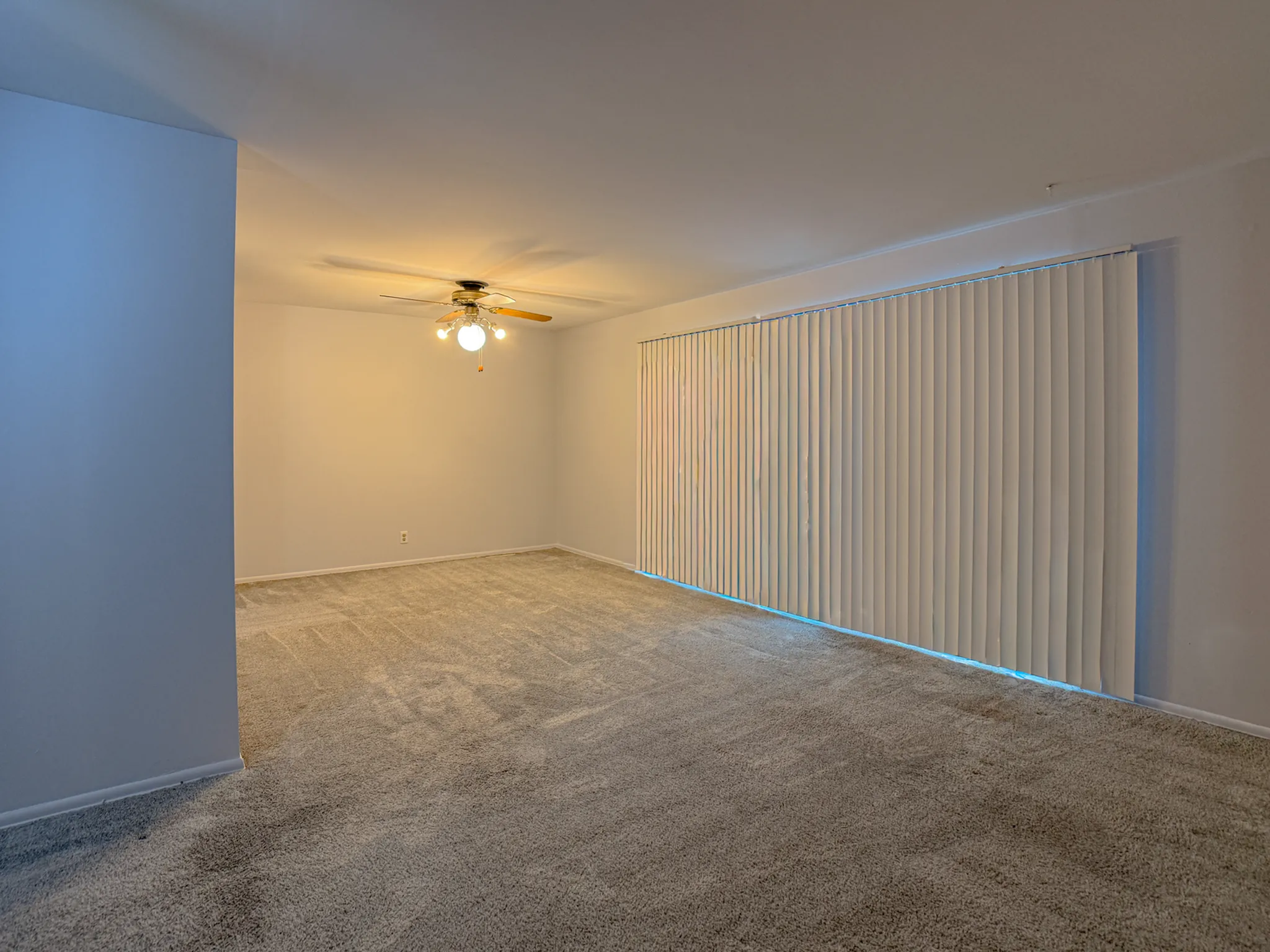 Spacious, carpeted living area with large window and ceiling fan, perfect for cozy gatherings.