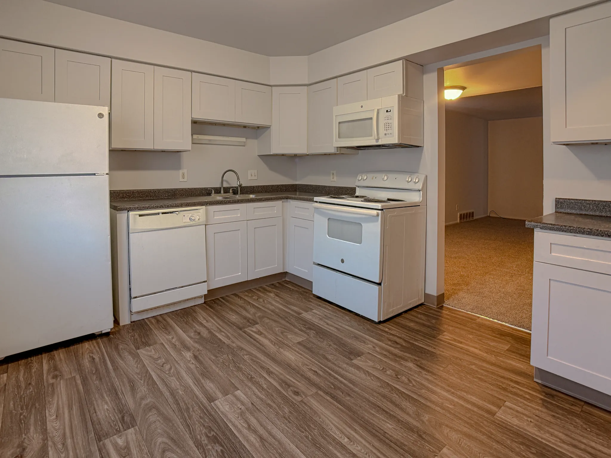 Modern kitchen with sleek white cabinets, stainless steel appliances, and elegant wood flooring, flowing into a cozy carpeted dining area.