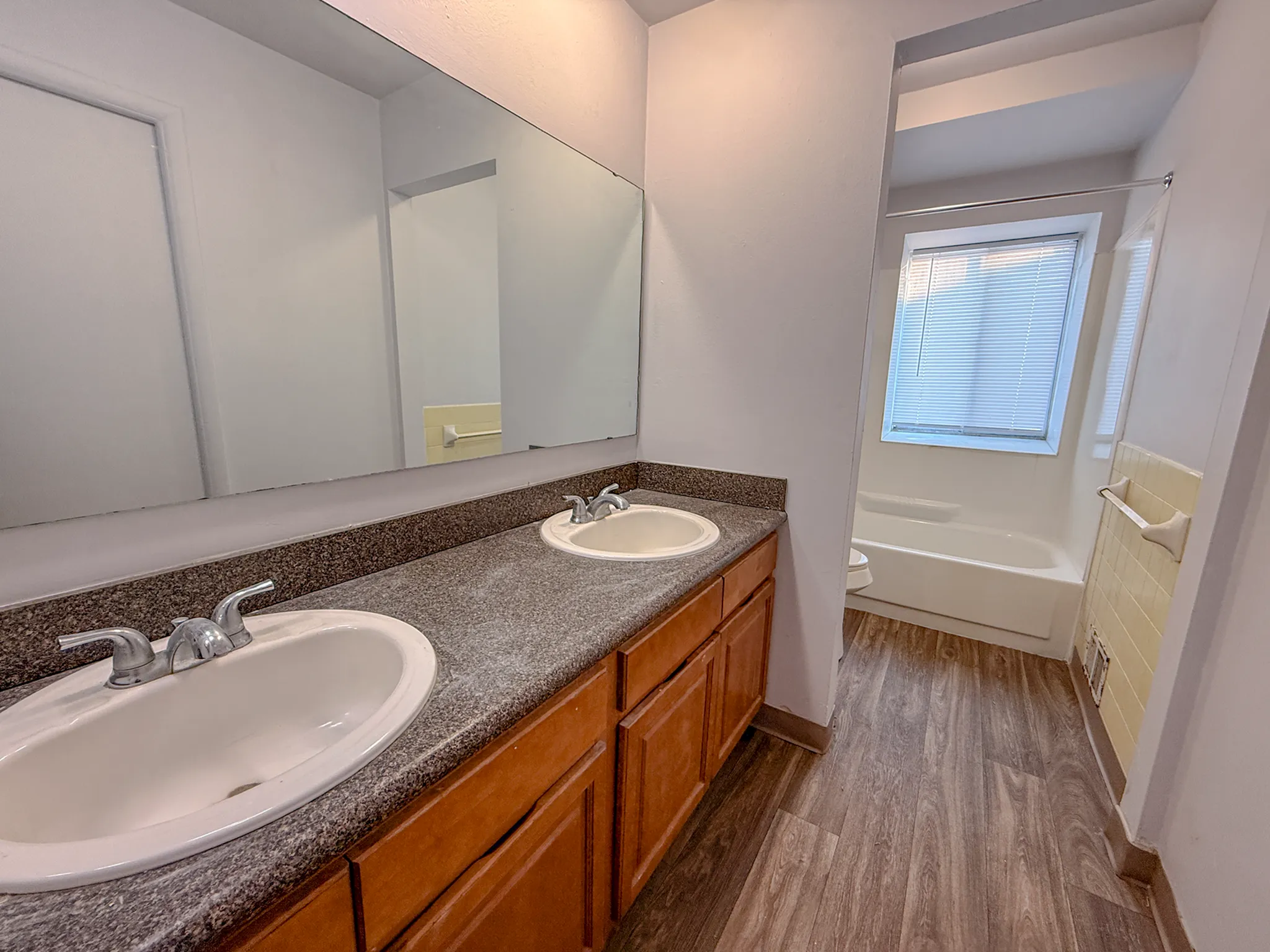 Bright bathroom with dual sinks, dark countertops, large mirror, and inviting bathtub, featuring elegant wood flooring.