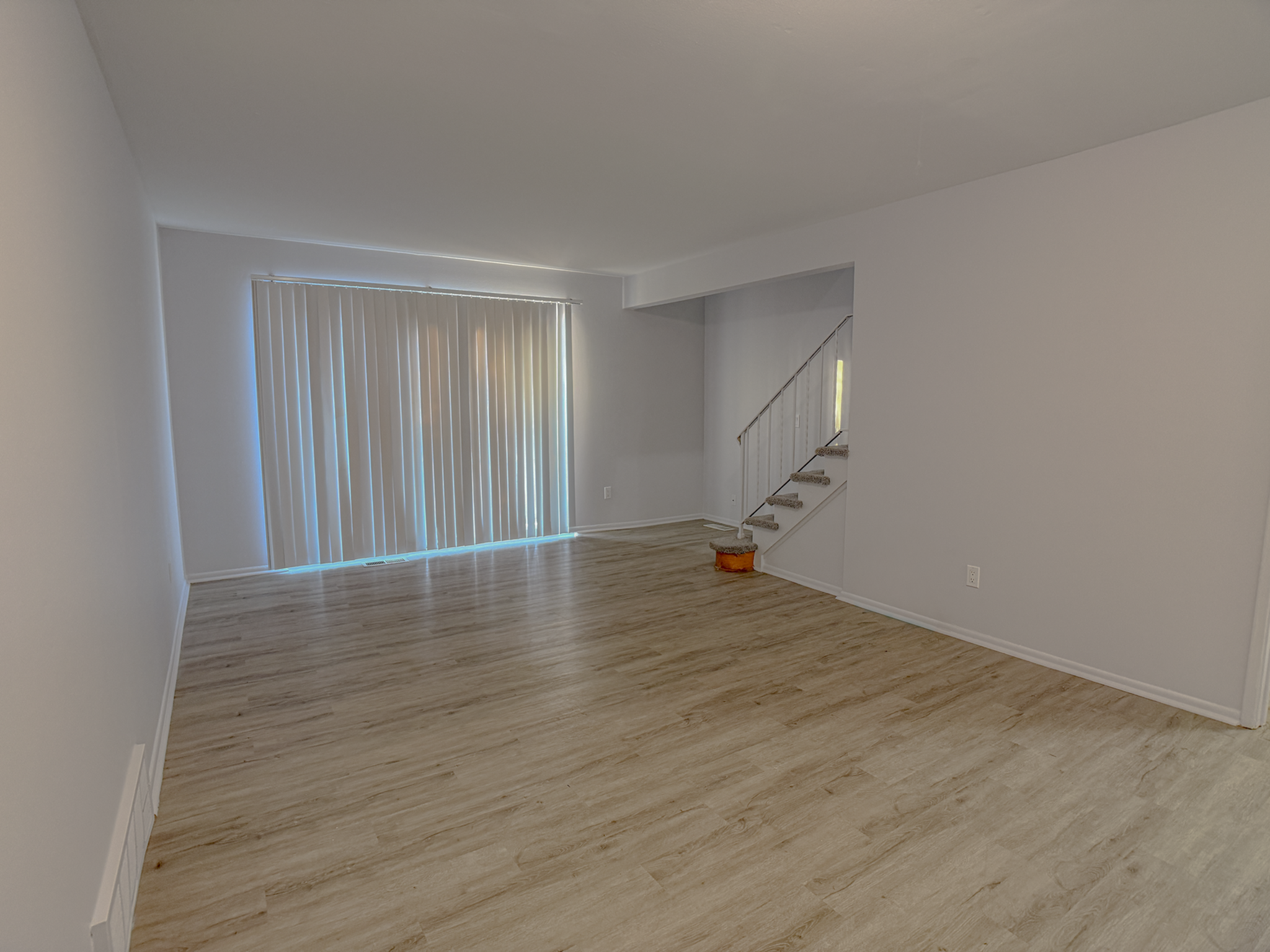 Bright and spacious living area with elegant wooden floors and modern staircase, complemented by expansive window blinds for ample natural light.