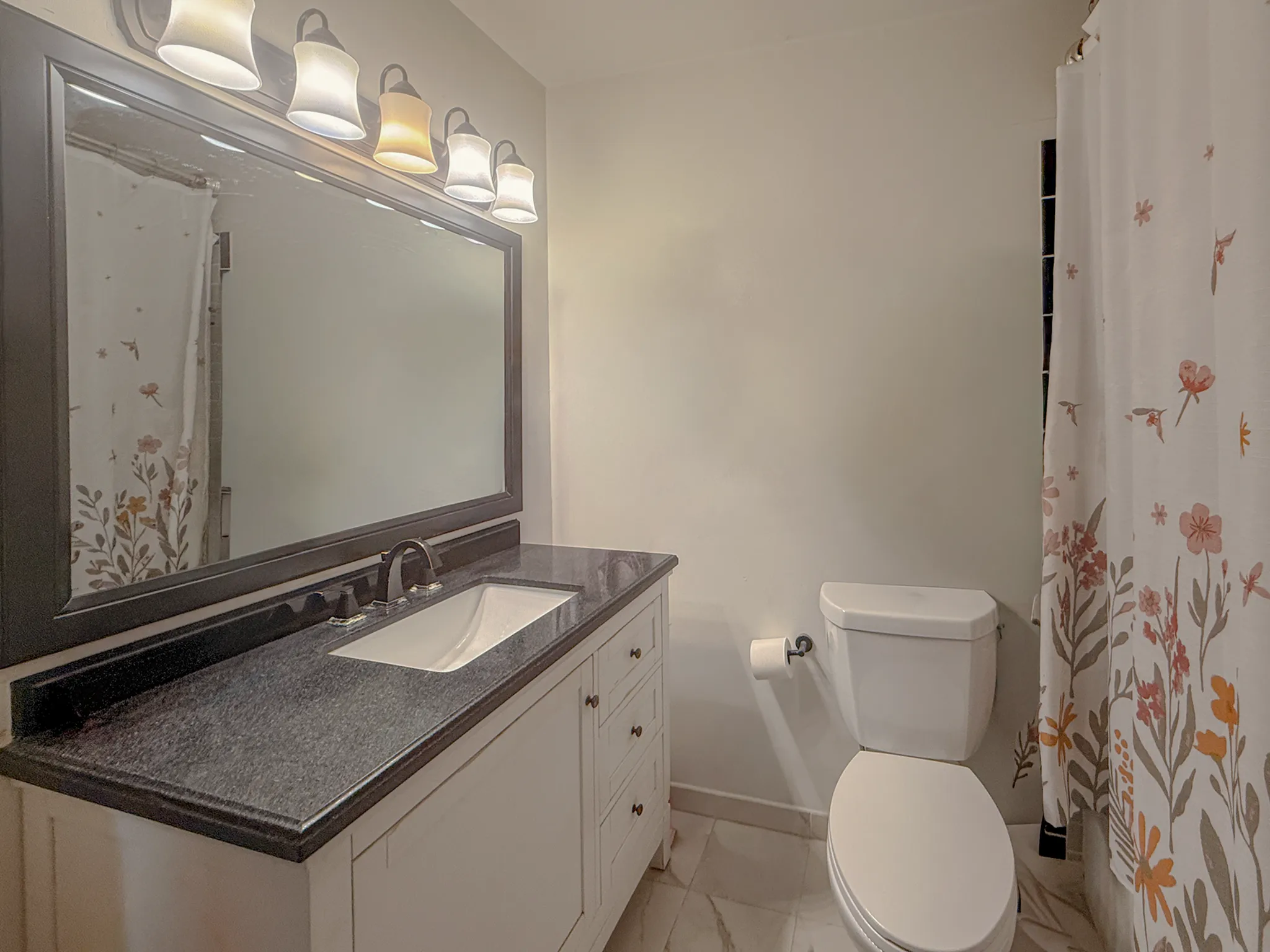 Charming bathroom featuring elegant lighting, a sleek black countertop, large mirror, and floral-patterned shower curtain.