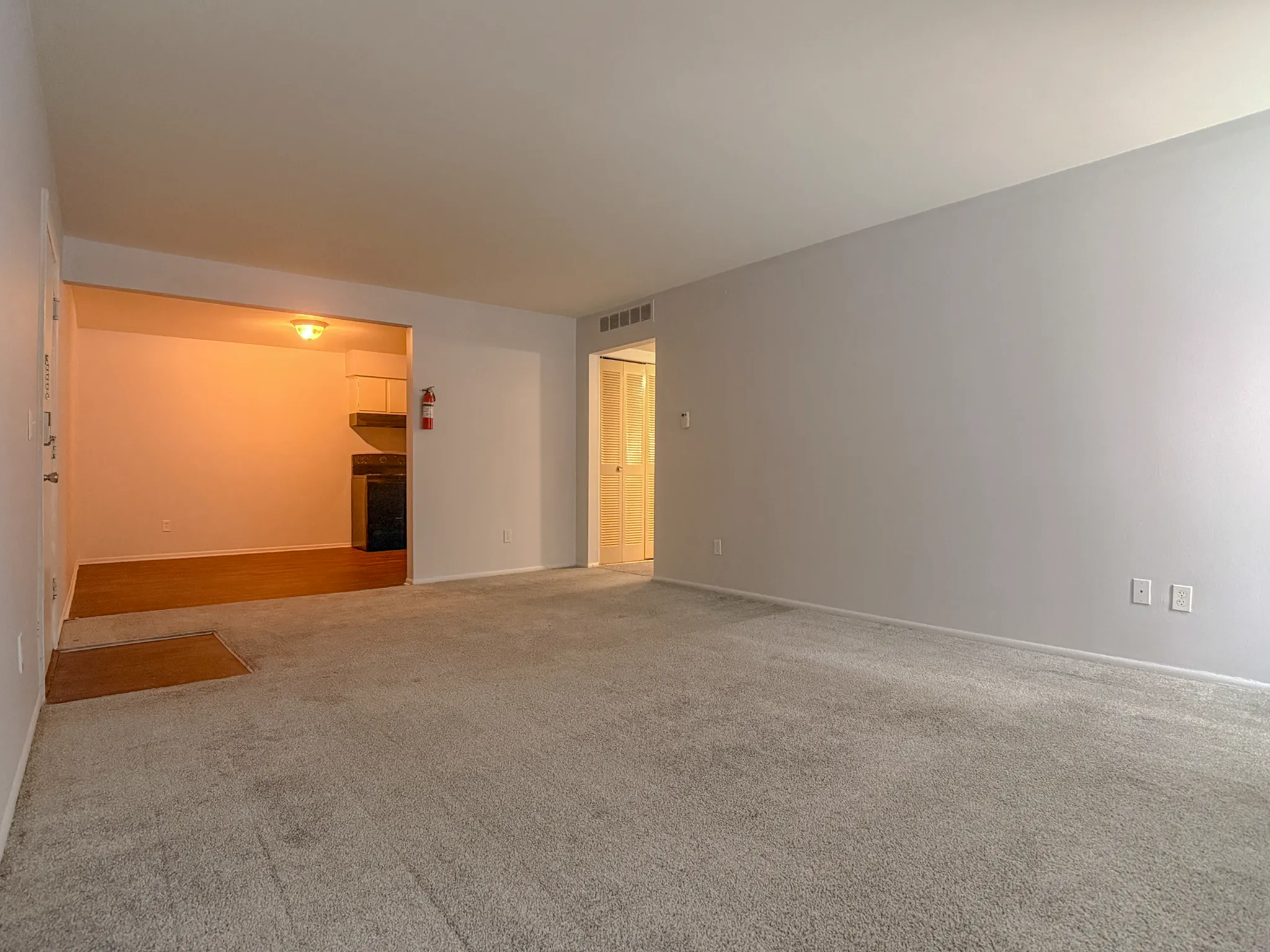 Spacious living area with cozy carpeting, adjacent to a warm lit kitchen, perfect for comfortable apartment living.