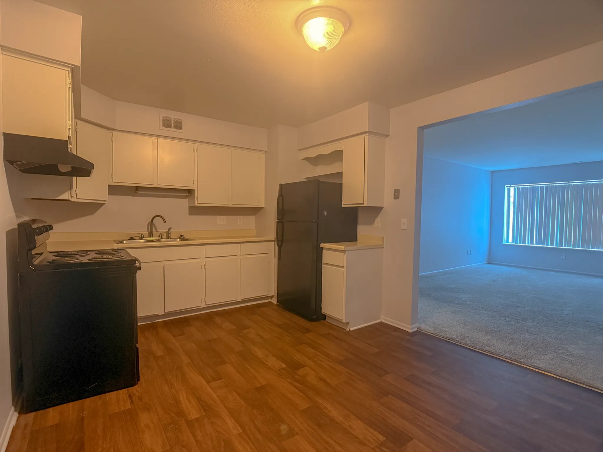 Inviting apartment kitchen with warm wood flooring, sleek black appliances, and a view into a spacious, well-lit living room.