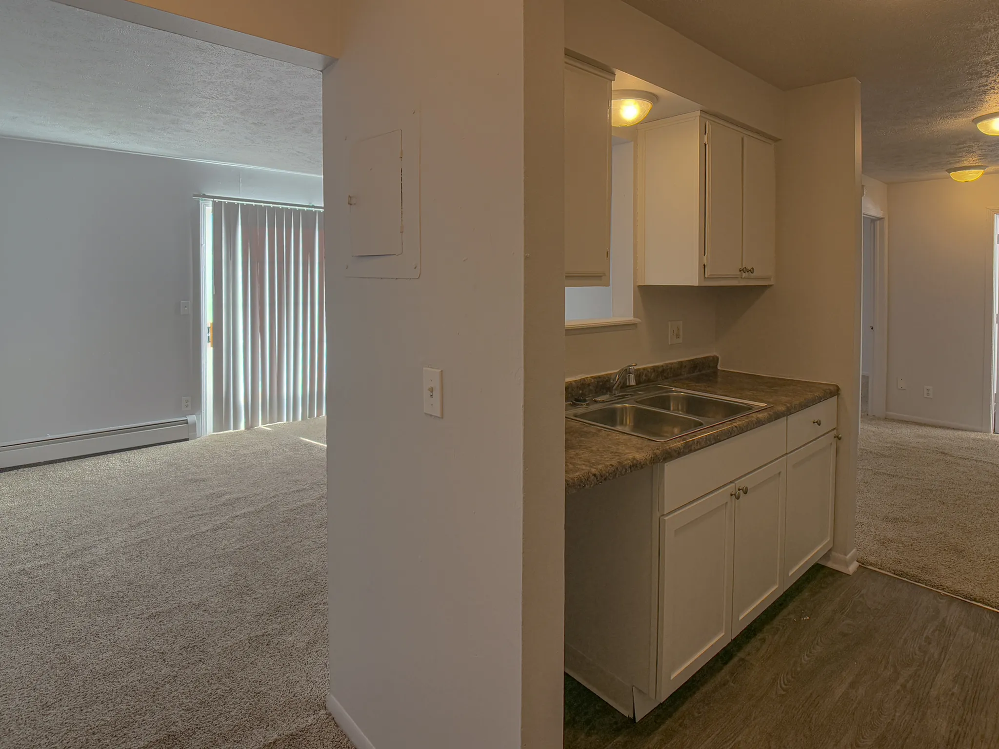 Inviting apartment featuring a cozy kitchen with modern cabinetry, leading to a sunlit living area with plush carpeting and sliding glass doors.
