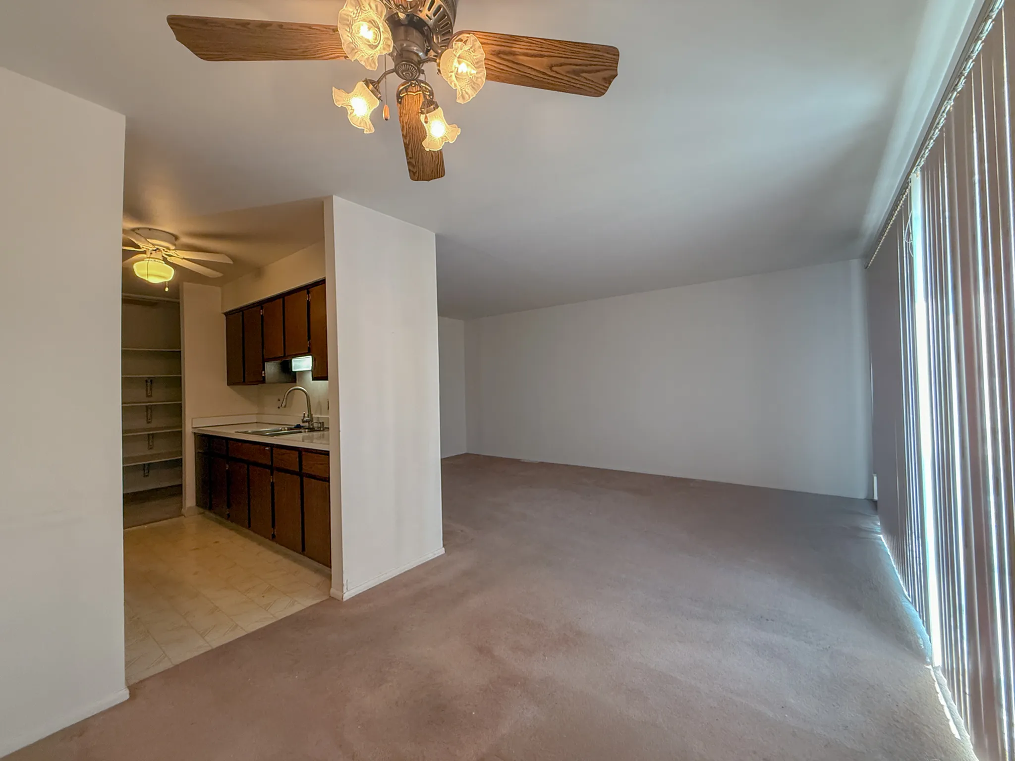 Bright and open living space with plush carpet and a ceiling fan, adjacent to a cozy kitchen with ample storage and wooden cabinetry.