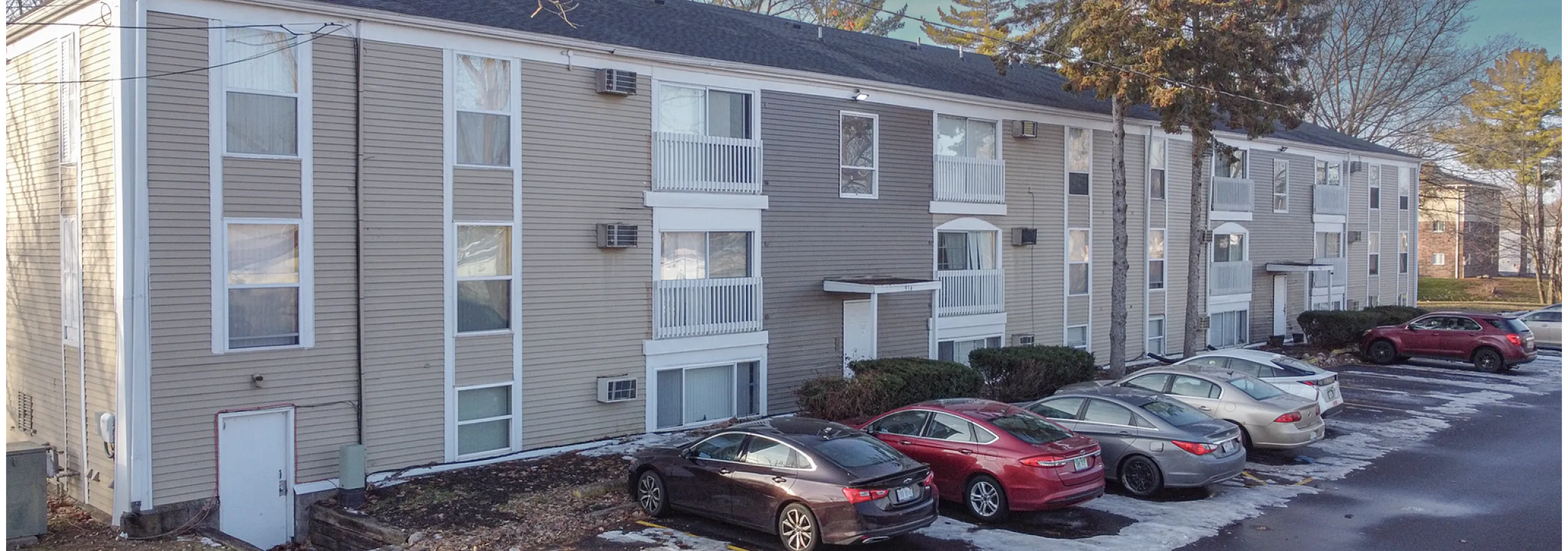 Charming three-story apartment building with ample parking, surrounded by lush greenery and tall trees.