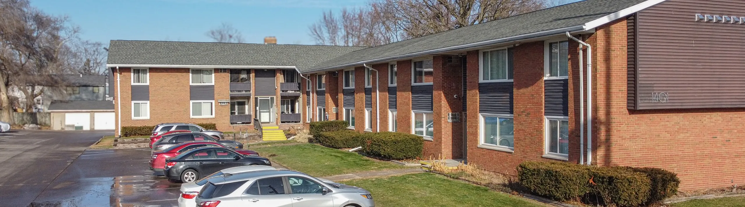 Charming brick apartment building with ample parking and well-maintained landscaping on a sunny day.