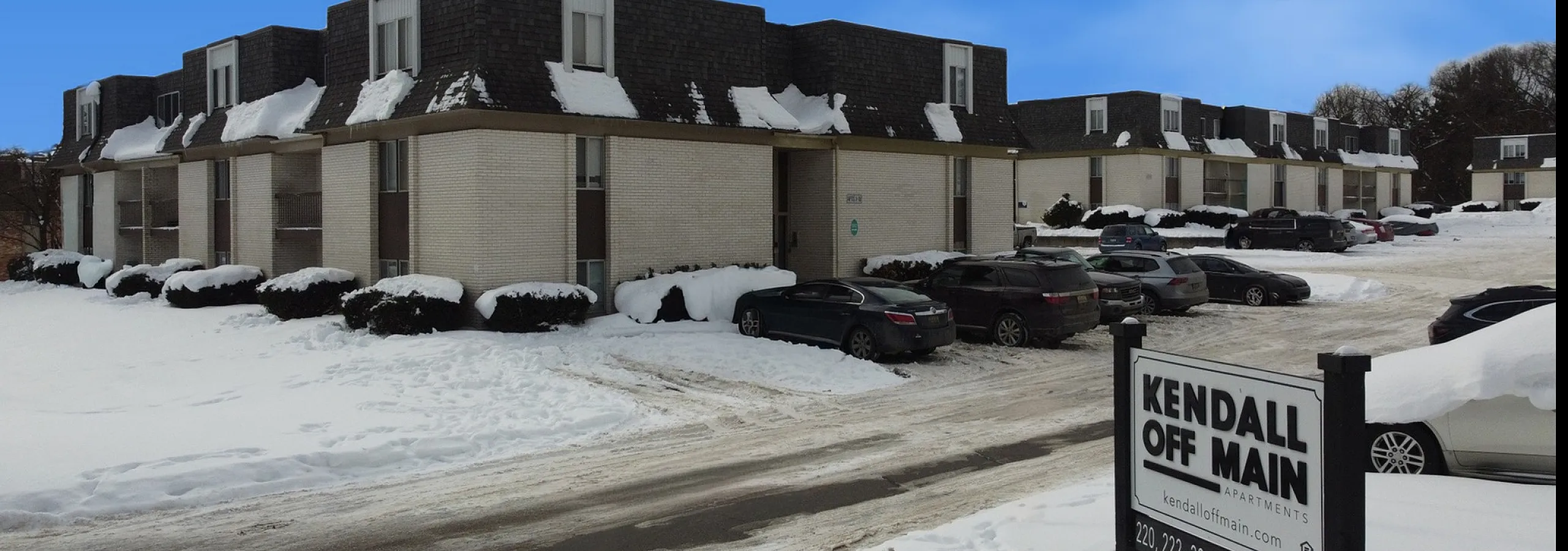 Charming winter view of Kendall Off Main Apartments with snow-covered landscape and ample parking.