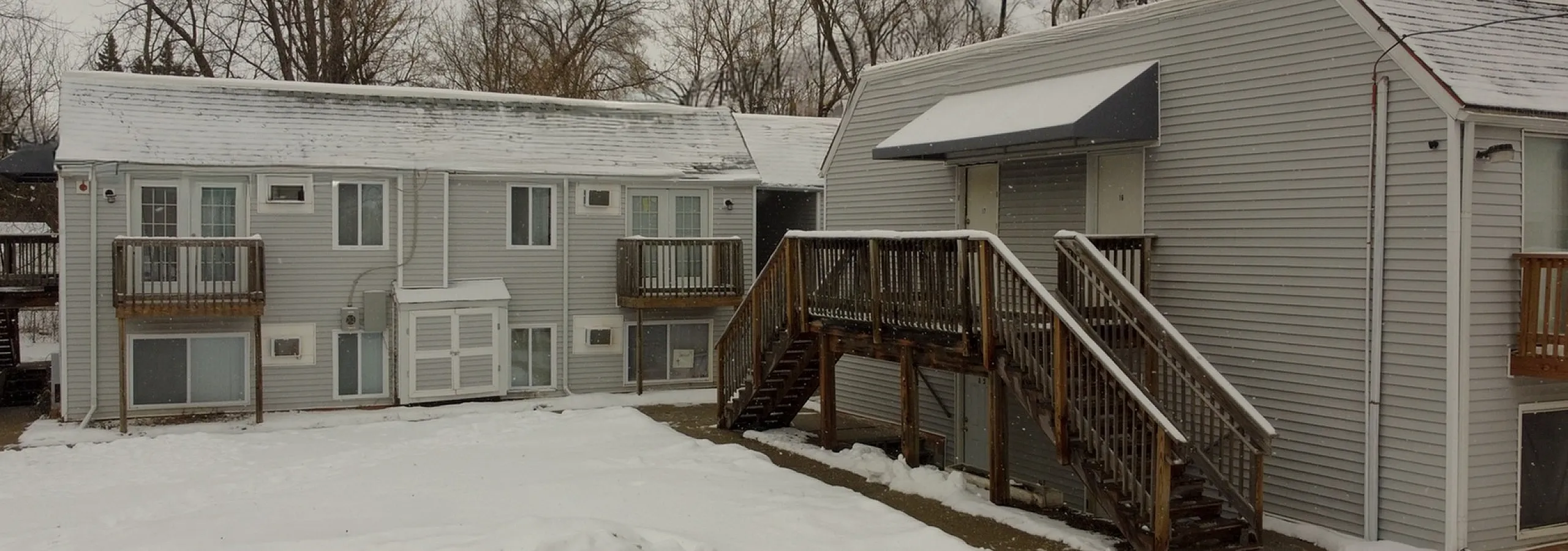 Charming snowy exterior view of a modern apartment building with wooden balconies and staircases, surrounded by serene winter trees.