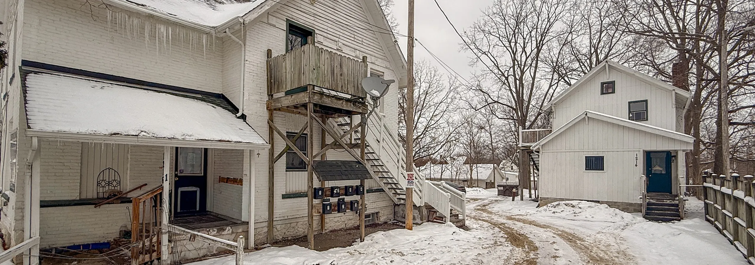 Charming winter scene of a rustic white building with a cozy balcony, surrounded by snow and framed by bare trees.
