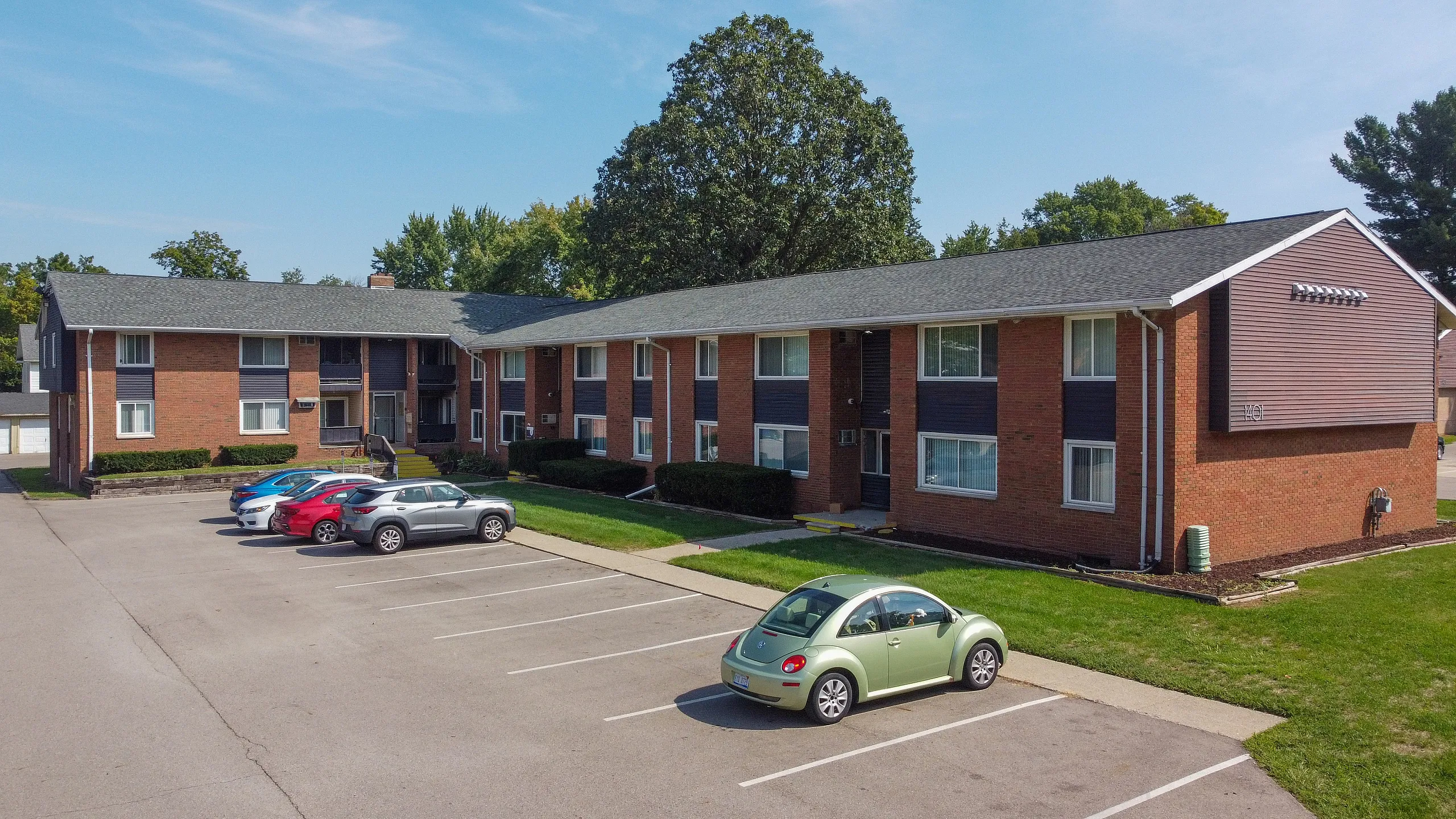 Charming brick apartment complex with ample parking, surrounded by lush greenery and blue skies, offering a welcoming community atmosphere.