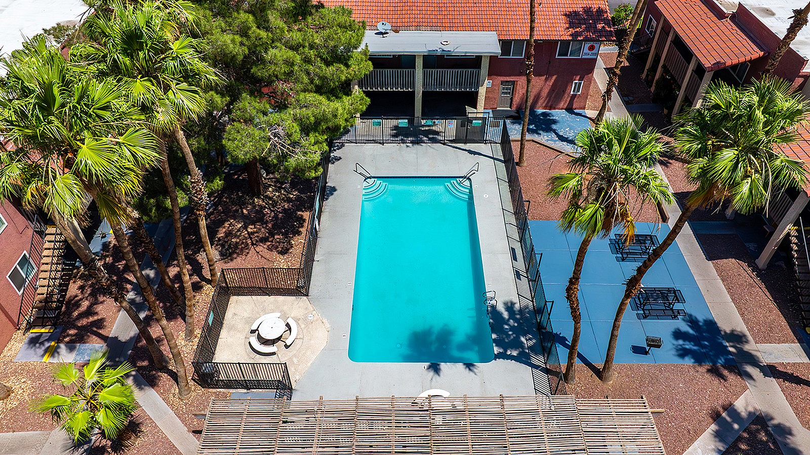 Inviting aerial view of a vibrant apartment complex featuring a sparkling swimming pool surrounded by lush palm trees and charming red-tiled roofs.