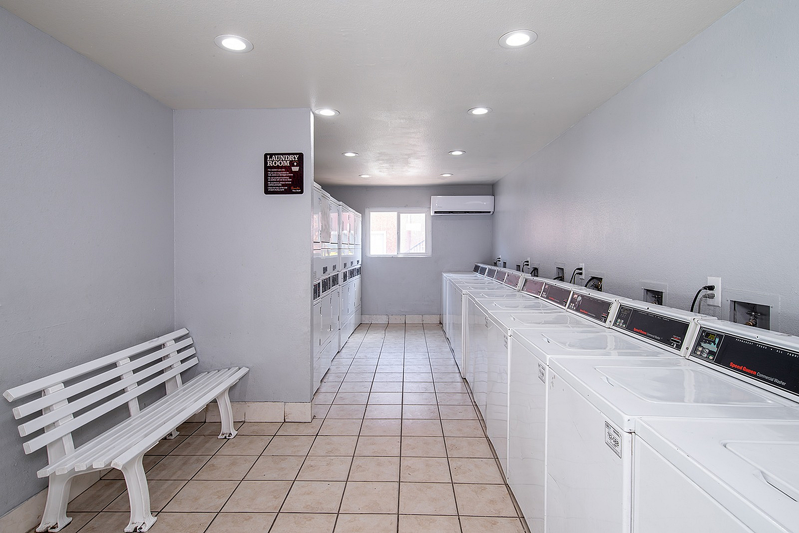 Modern and bright communal laundry room featuring multiple washing machines, tiled floors, and a cozy white bench under soft lighting.