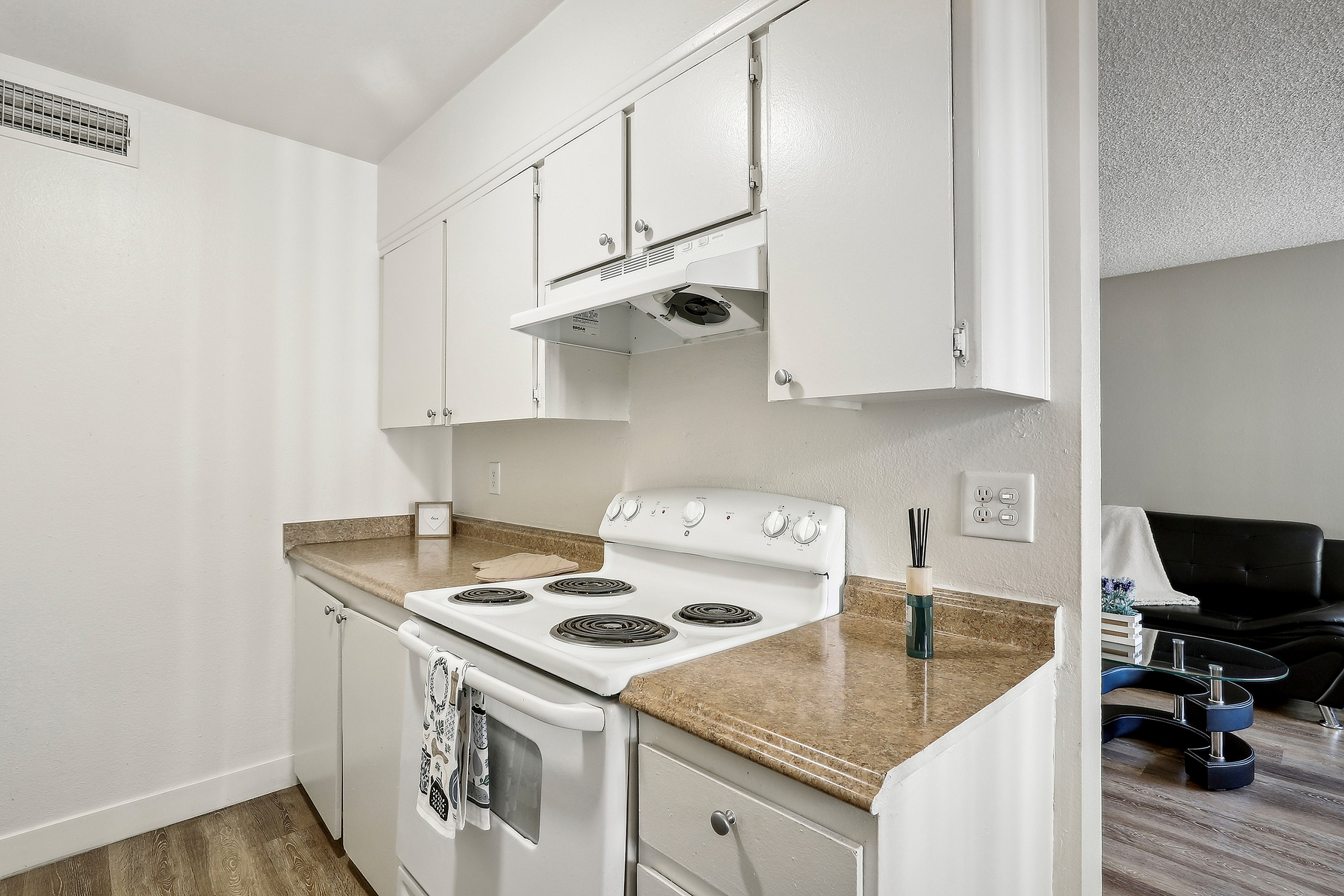 Charming compact kitchen featuring sleek white cabinets, an electric stove, and warm brown countertops, seamlessly extending to a cozy living area.