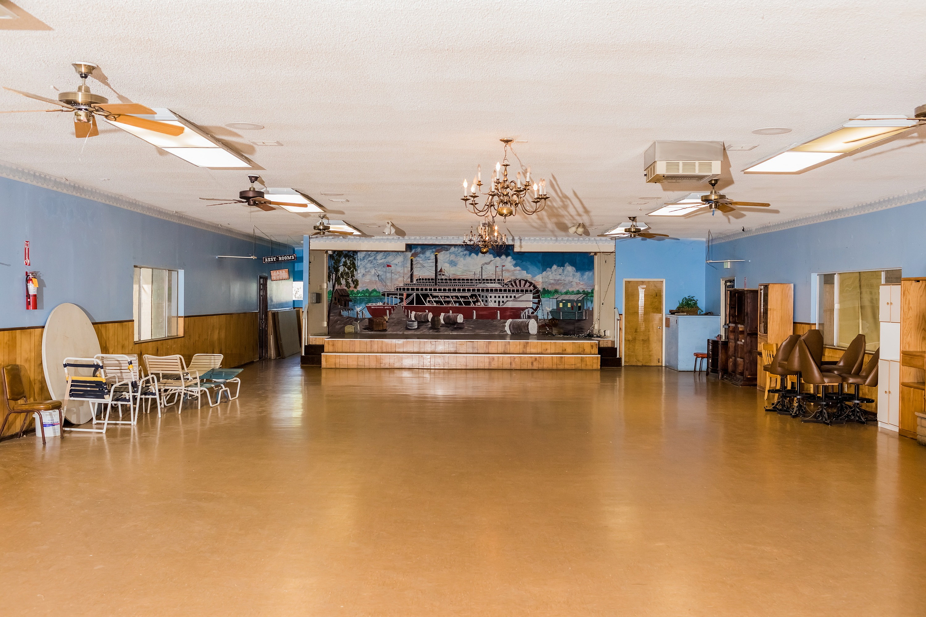 Spacious community hall with elegant chandeliers, ceiling fans, and a charming mural backdrop, perfect for events and gatherings.