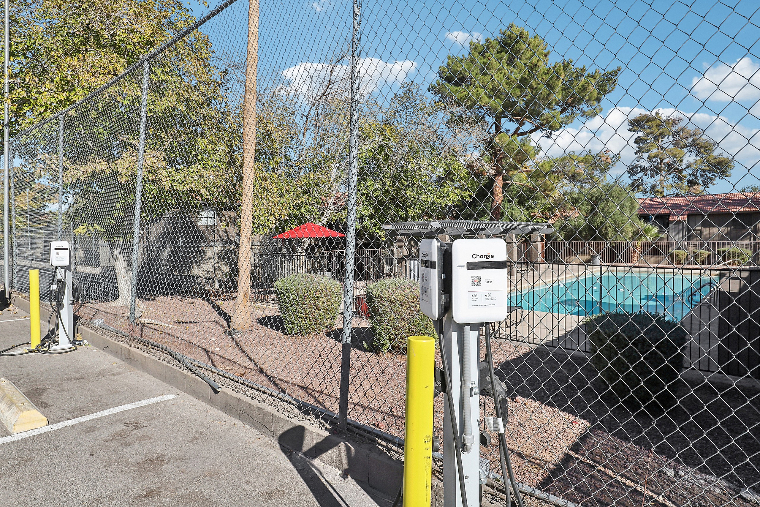 Modern electric vehicle charging station beside a serene swimming pool enclosure, surrounded by lush greenery under a clear blue sky.