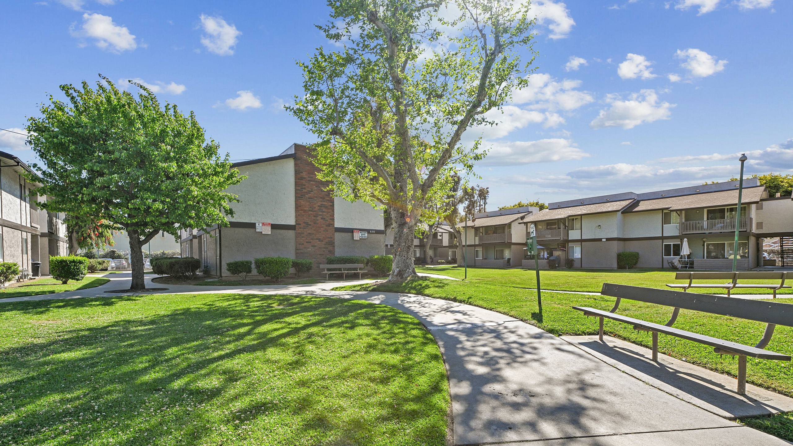 Charming apartment complex with lush green lawns, mature trees, and inviting benches, set under a bright blue sky.