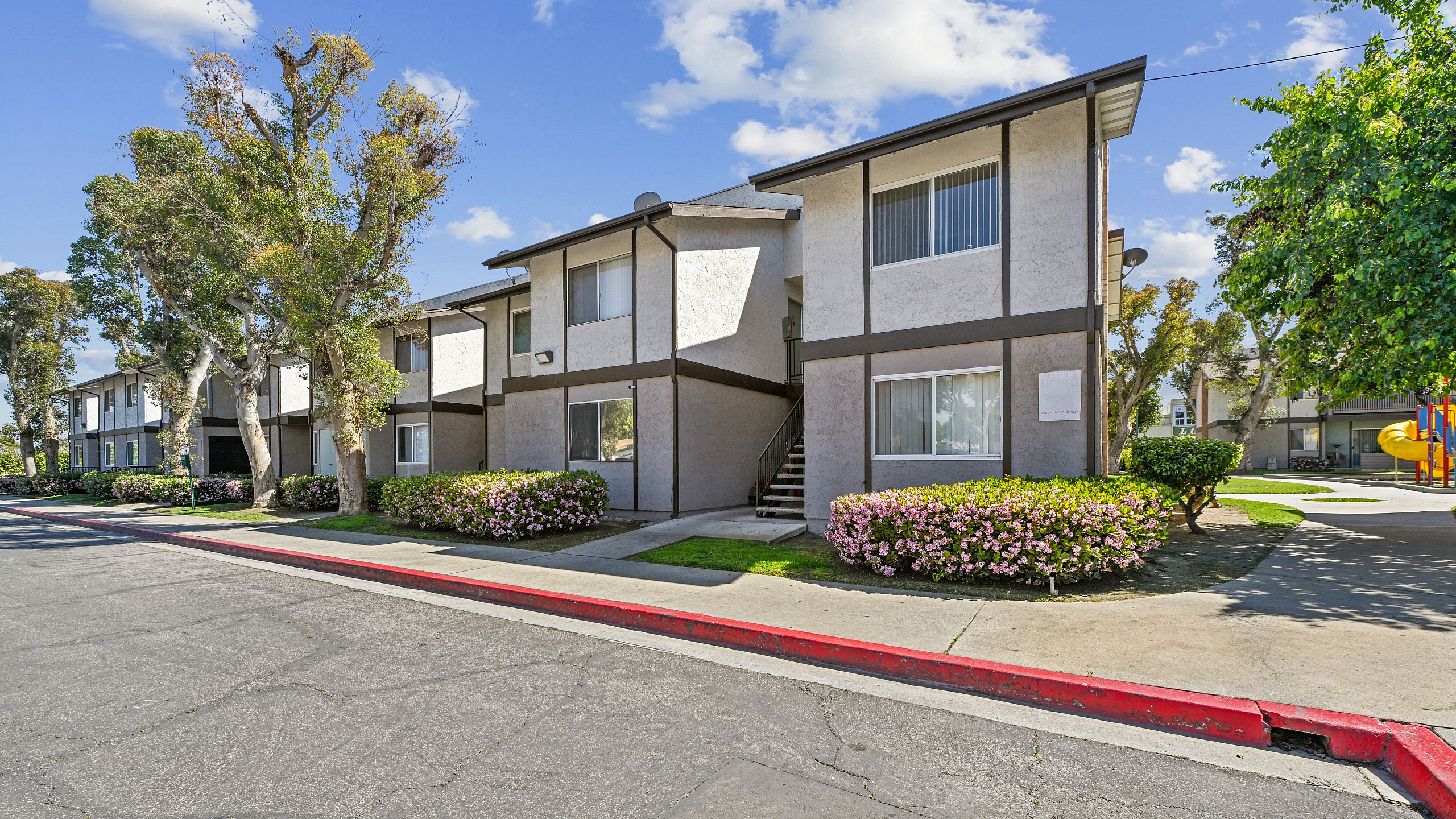 Charming apartment complex with manicured landscaping, vibrant flowers, and mature trees under a bright blue sky.