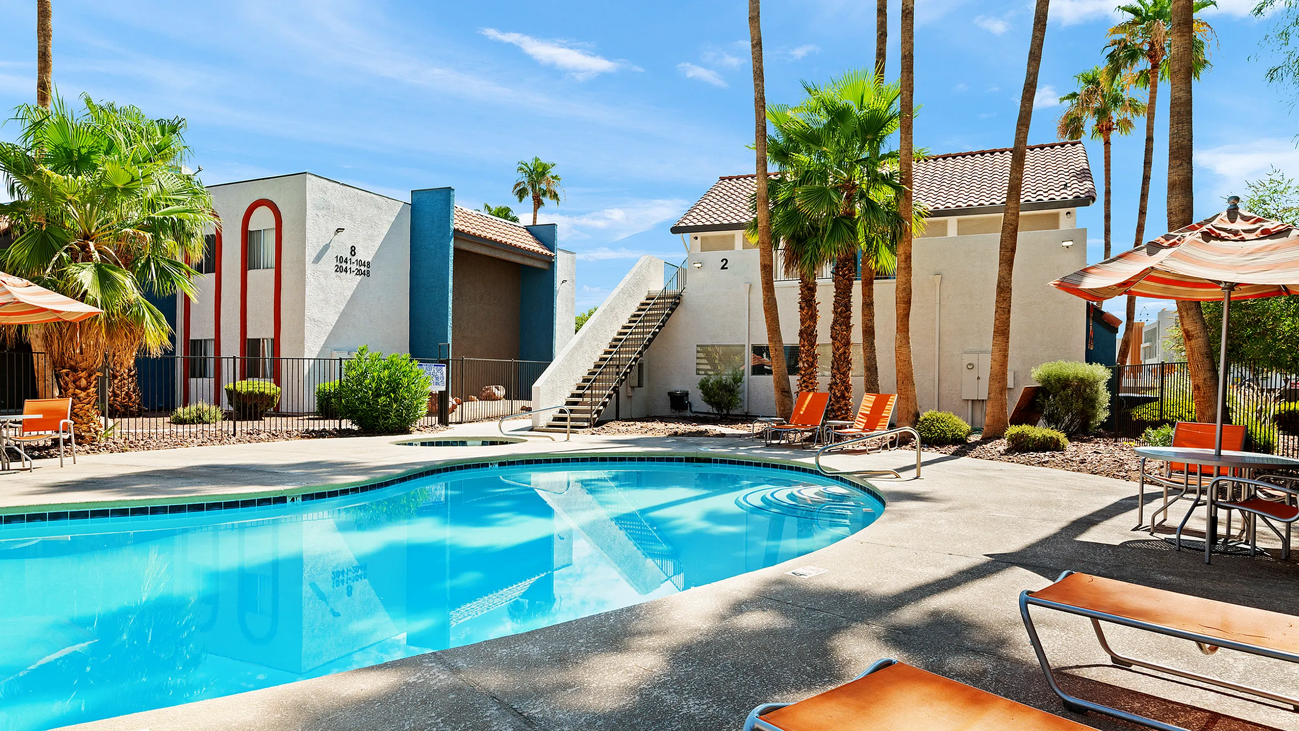 Inviting apartment complex pool area with colorful loungers and palm trees, under a bright blue sky.