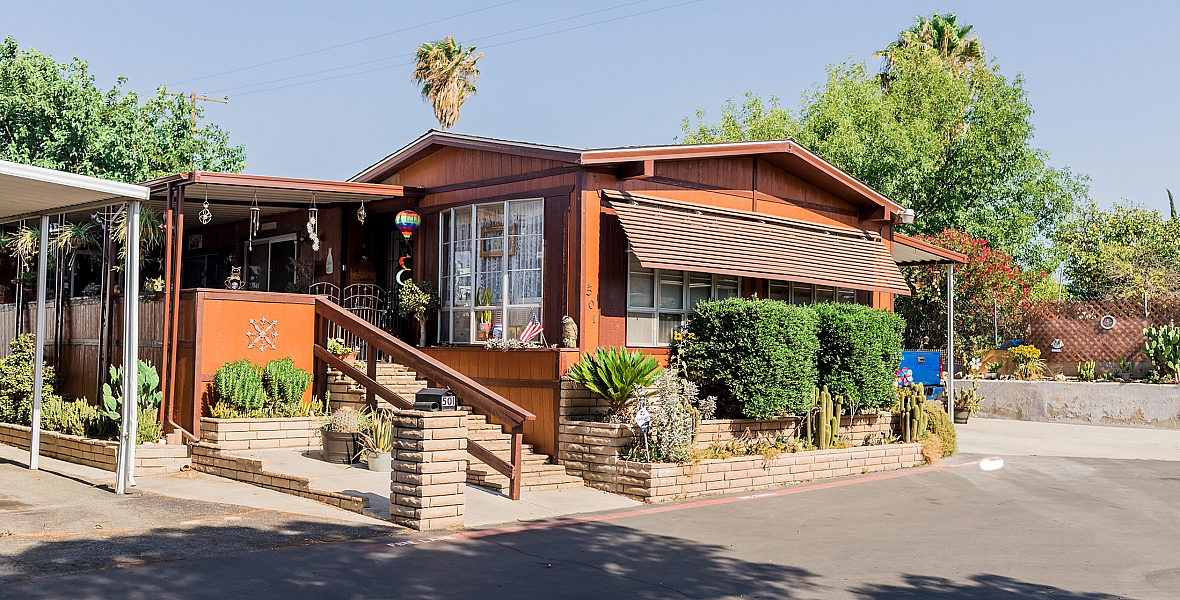 Charming mobile home with a wooden exterior, surrounded by lush greenery and a beautifully landscaped front yard, ideal for cozy living.