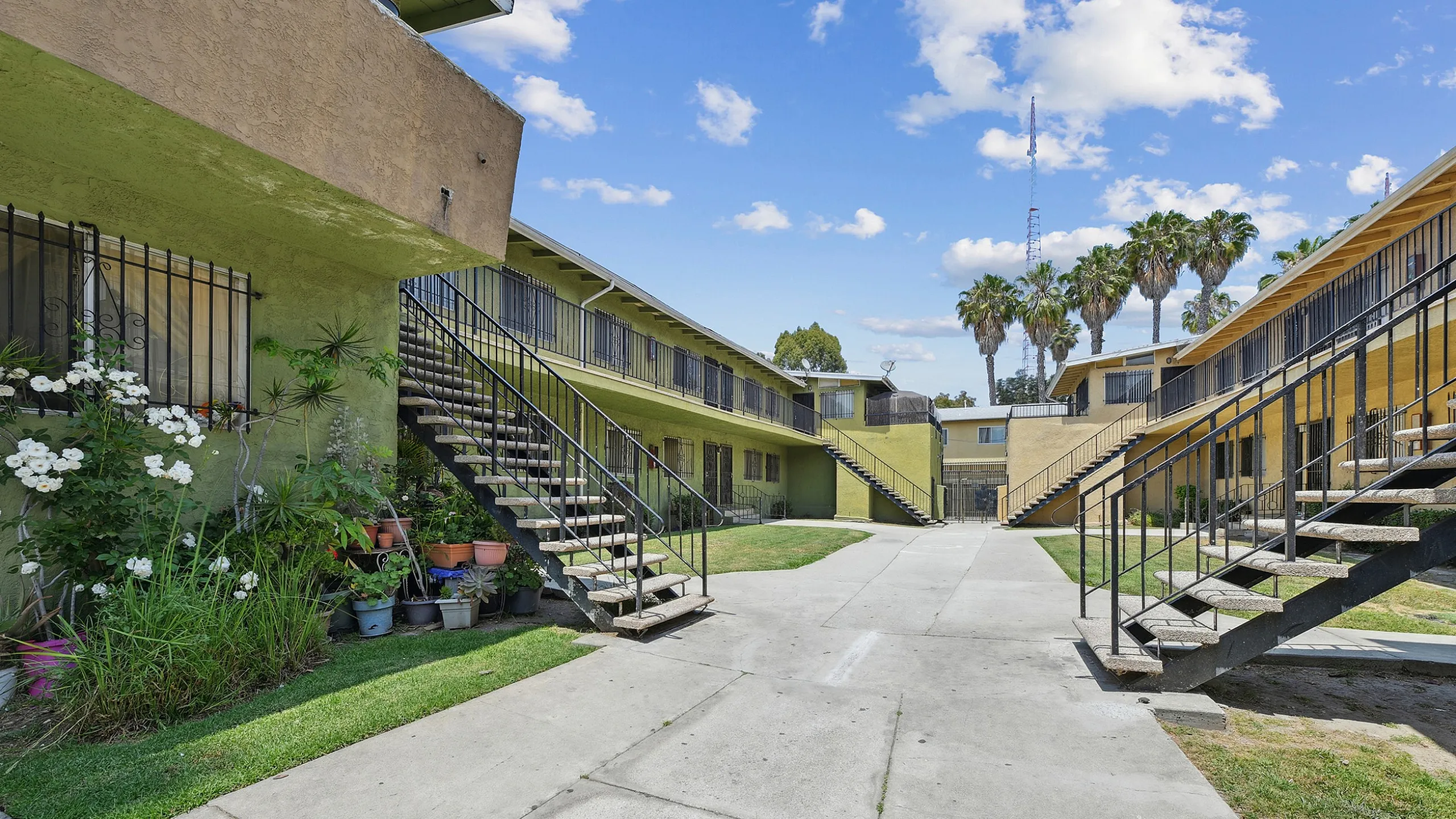 Charming apartment complex with lush greenery, vibrant flowers, and palm trees under a bright blue sky.