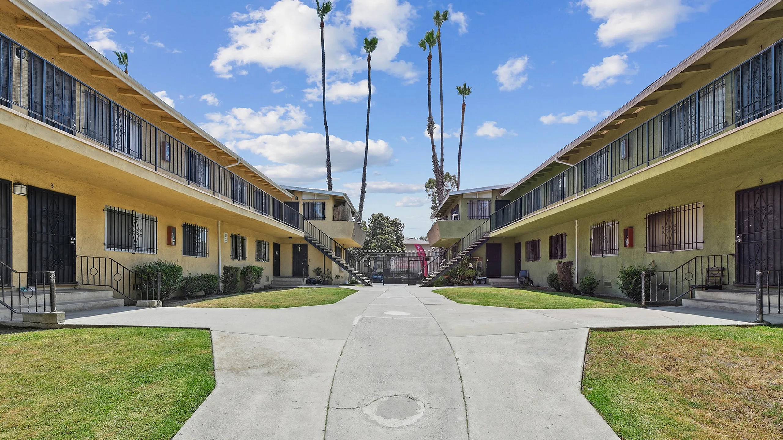 Charming apartment complex with two-story buildings, manicured lawns, and a welcoming walkway under a bright blue sky.