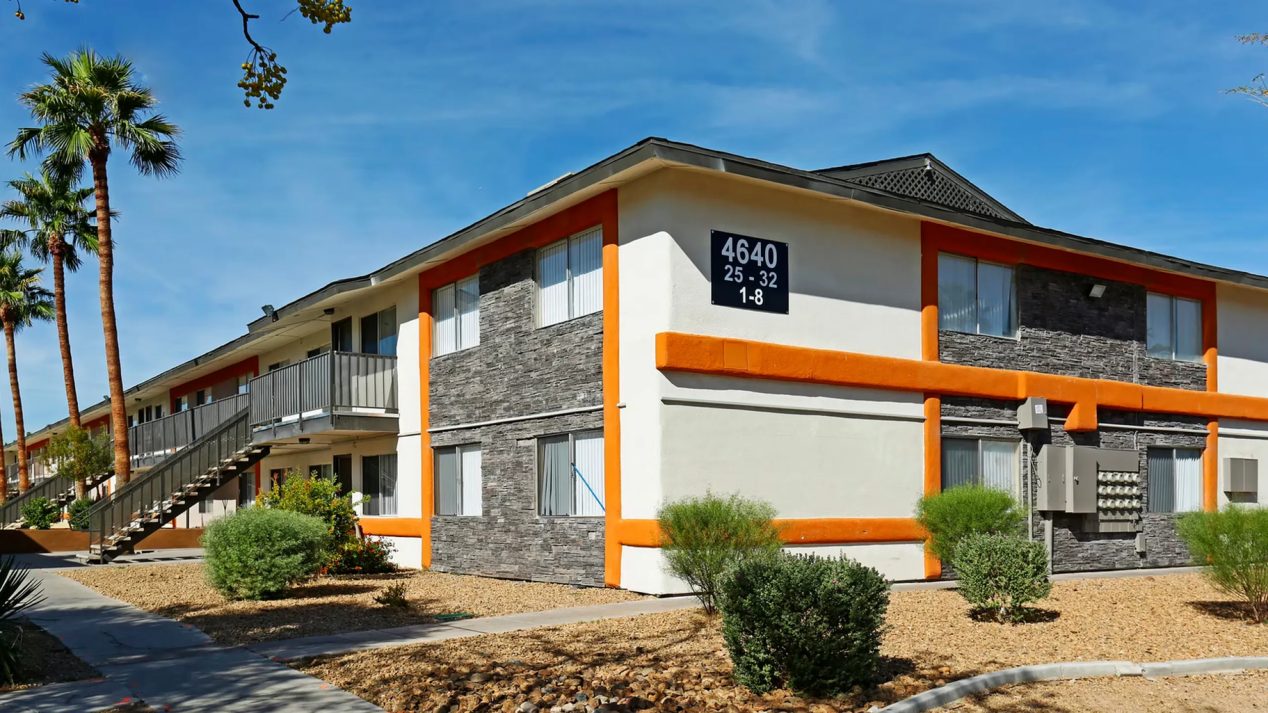 Modern two-story apartment building with stylish stone accents and vibrant orange trim nestled amidst desert landscaping, under a clear blue sky.