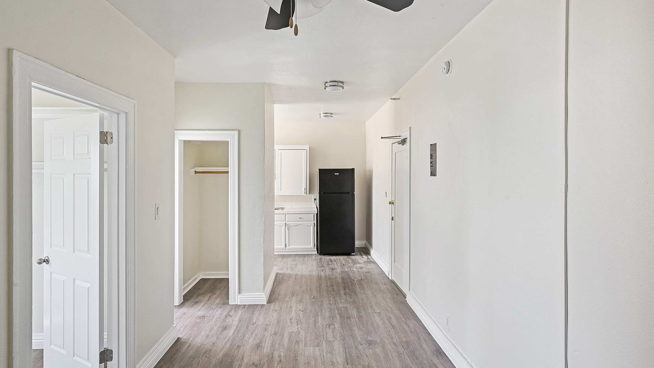 Bright and welcoming apartment hallway featuring sleek wood flooring, a modern ceiling fan, and a visible entrance to a cozy kitchen with white cabinets and a black refrigerator.