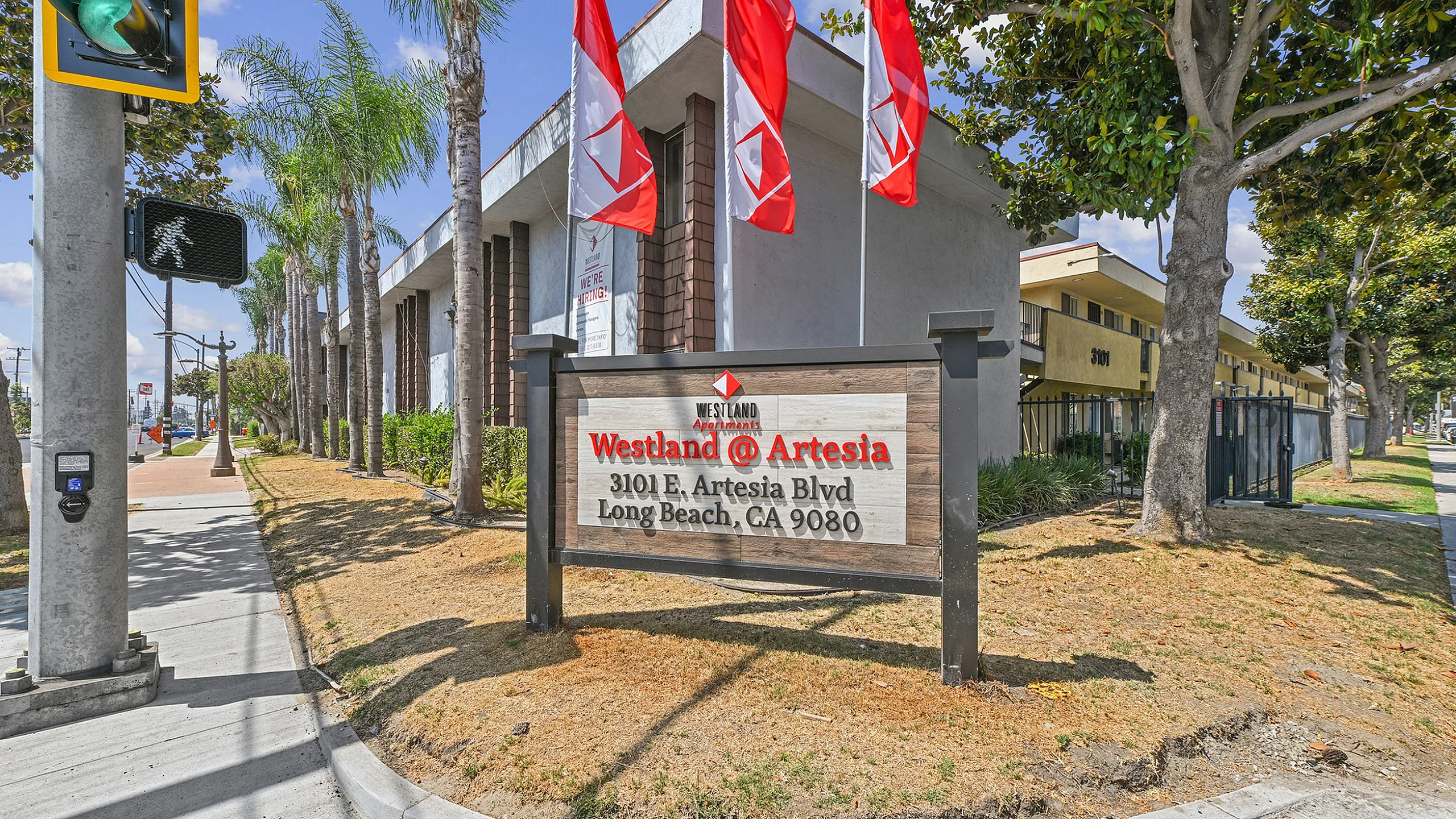 Inviting community entrance at Westland @ Artesia, featuring palm trees and vibrant flags in Long Beach.
