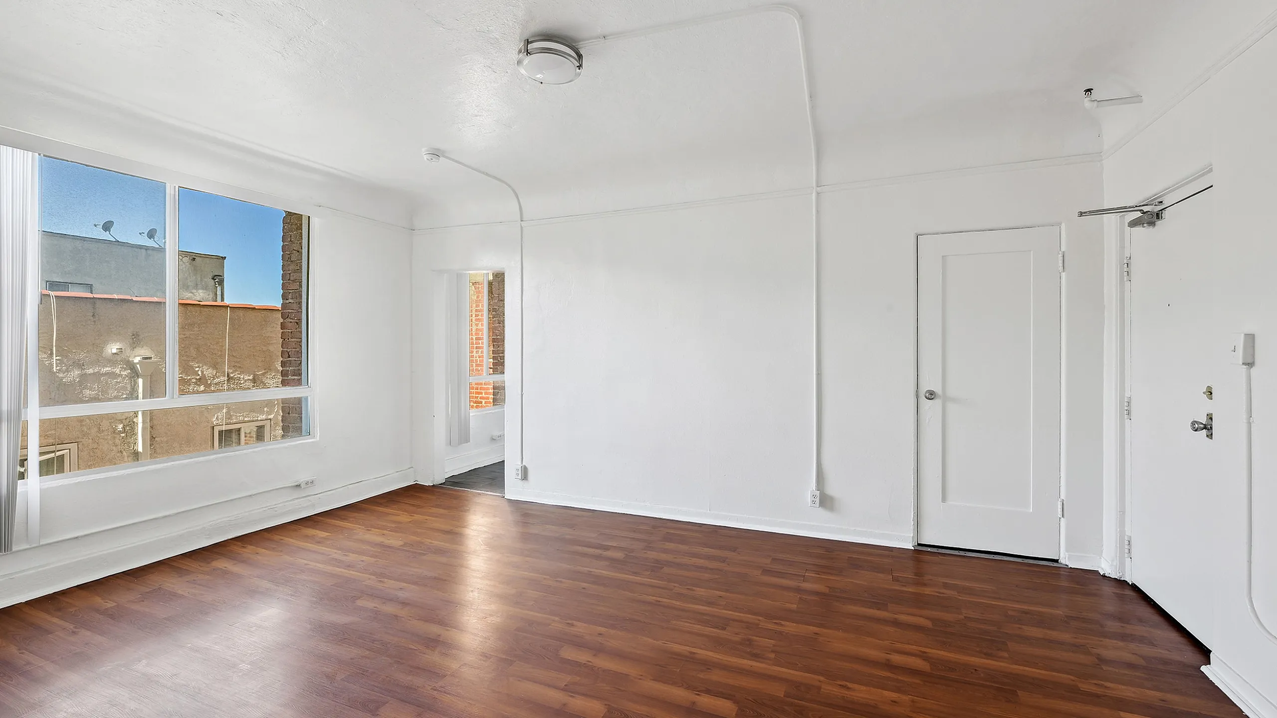Bright and spacious room with fresh white walls and rich wood flooring, featuring large windows inviting natural light and offering city views.