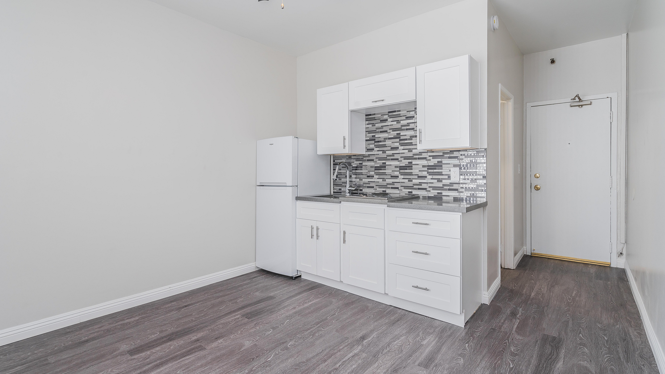Charming apartment kitchen with modern white cabinetry, sleek gray countertops, and stylish tile backsplash, complemented by elegant wood flooring and a ceiling fan.