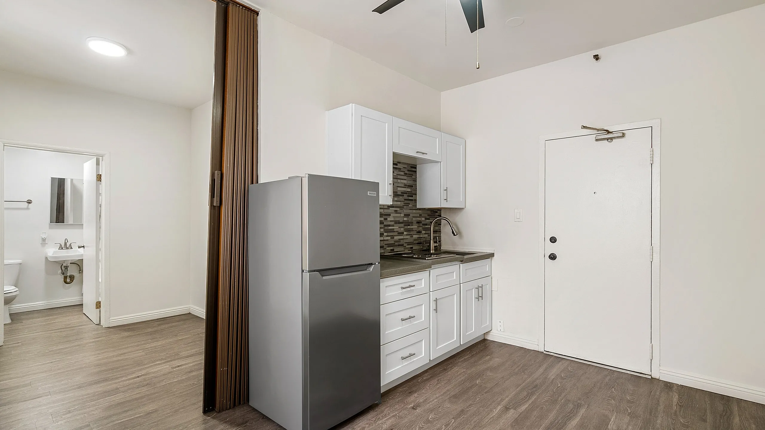 Modern apartment interior featuring a sleek kitchenette with white cabinetry and stainless steel fridge, complemented by elegant flooring and a convenient bathroom doorway.