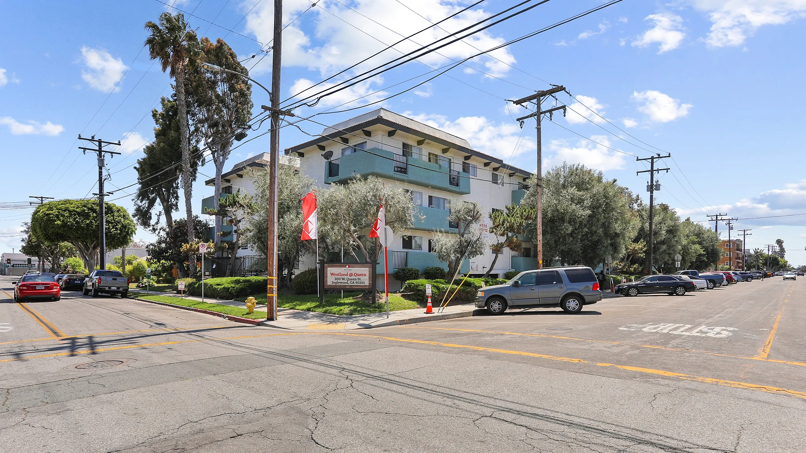 Charming multi-story apartment building with lush greenery and vibrant flags, set on a sunny suburban street corner.