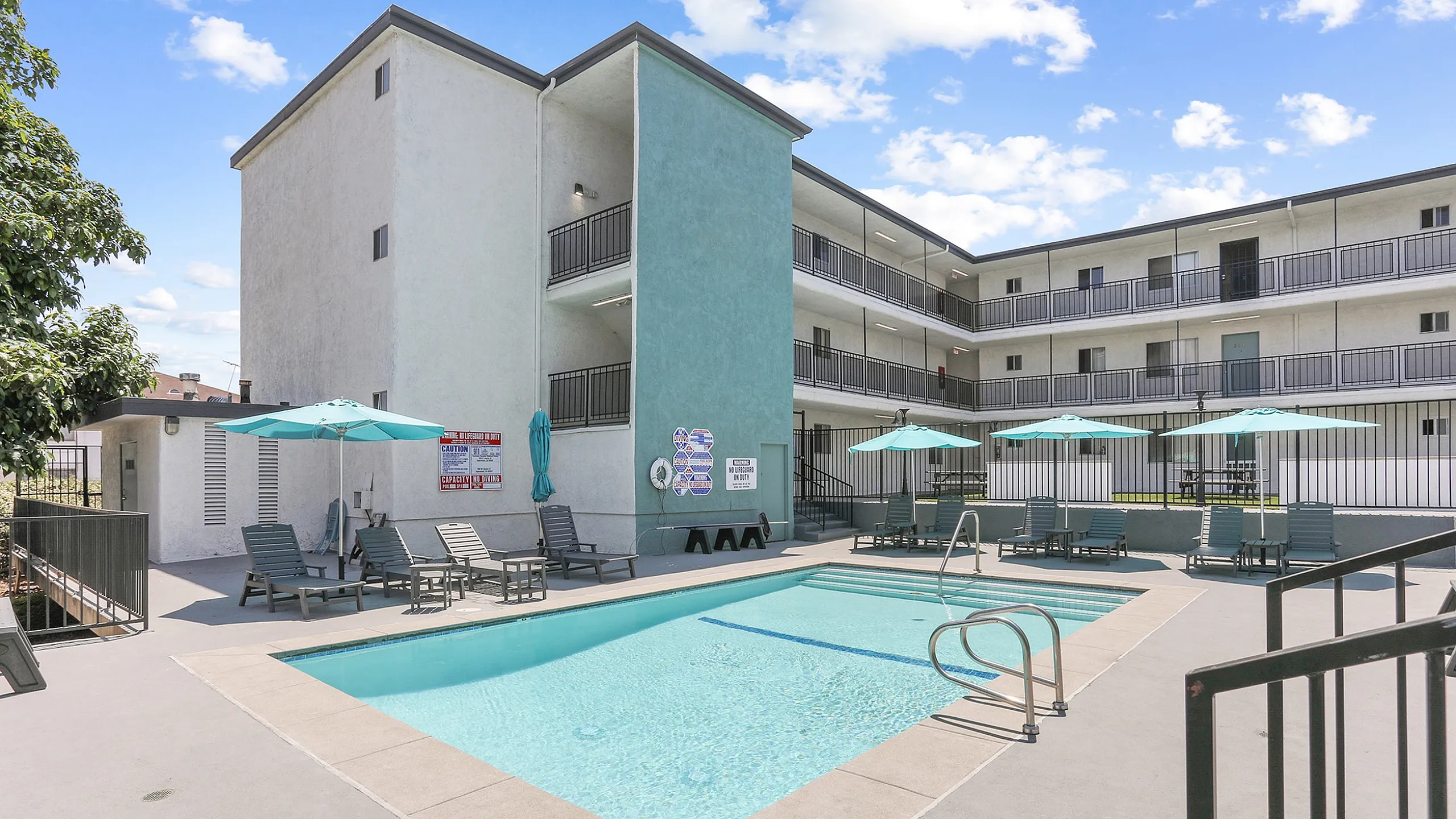 Charming apartment complex featuring a sunlit pool area with lounge chairs and umbrellas, surrounded by modern three-story buildings on a bright, clear day.