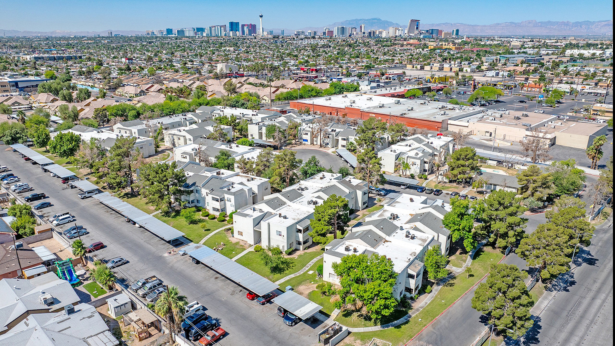 Aerial view of a vibrant apartment complex surrounded by lush greenery, parking spaces, and a bustling city backdrop.