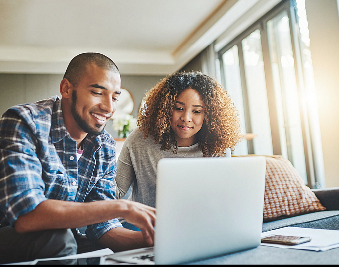 Happy couple using a laptop in a bright, modern living room with large windows and cozy decor.