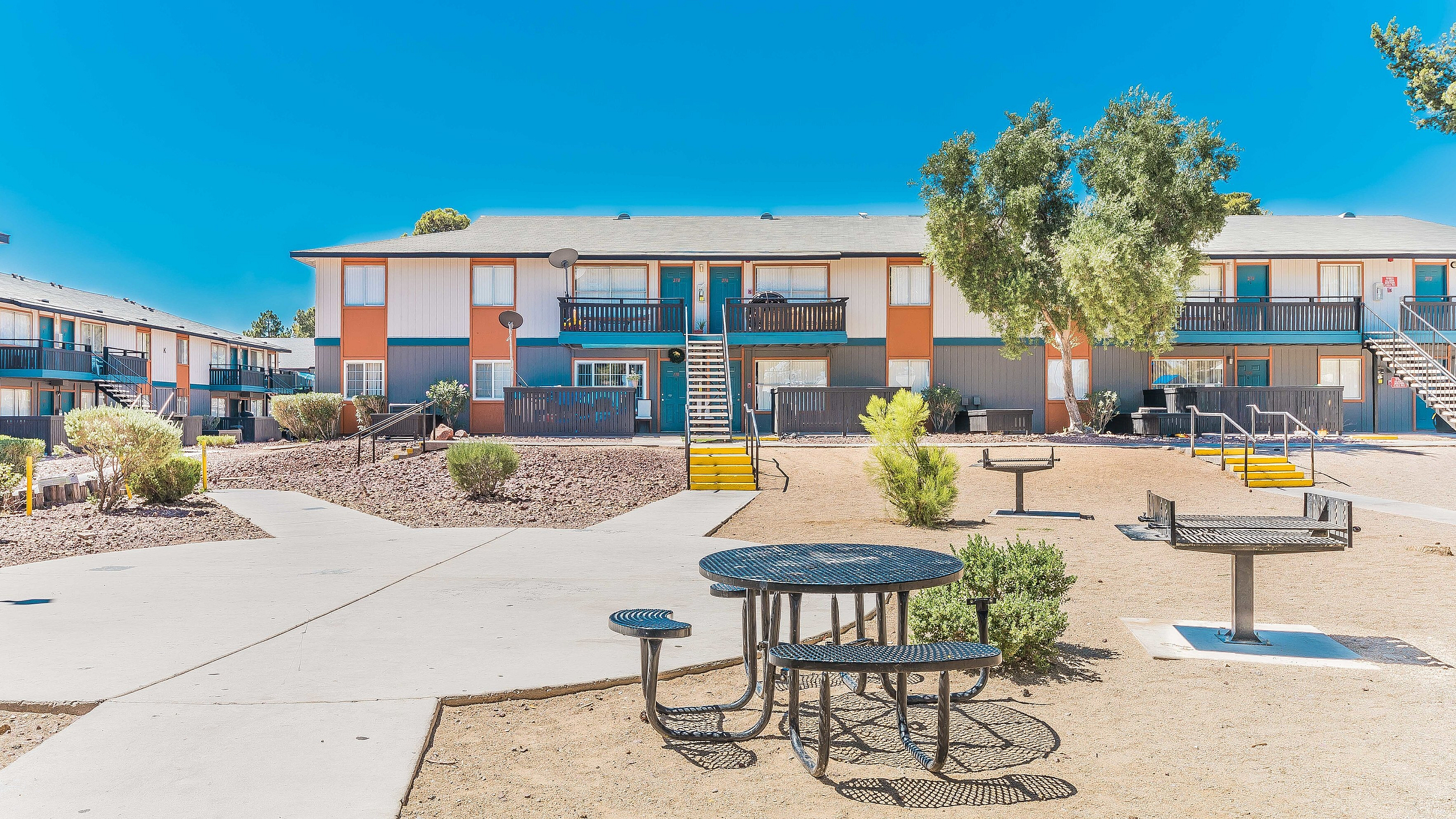 Bright, welcoming apartment complex with vibrant exterior, set against a clear blue sky, featuring well-maintained pathways and outdoor seating for a refreshing community experience.